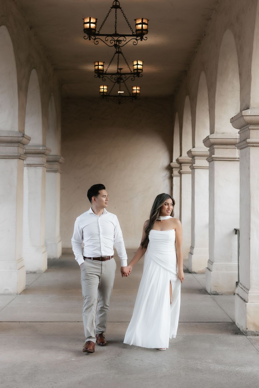 Couple holding hands walking under ornate chandelier through a beautiful arched corridor with tall columns.