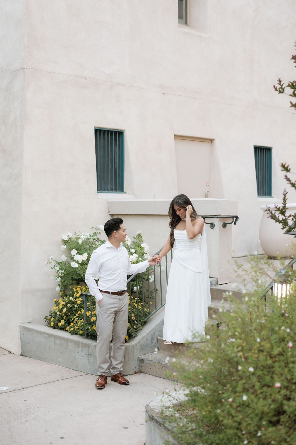 Couple in coordinating white outfits standing near steps with yellow flowers outside a building.