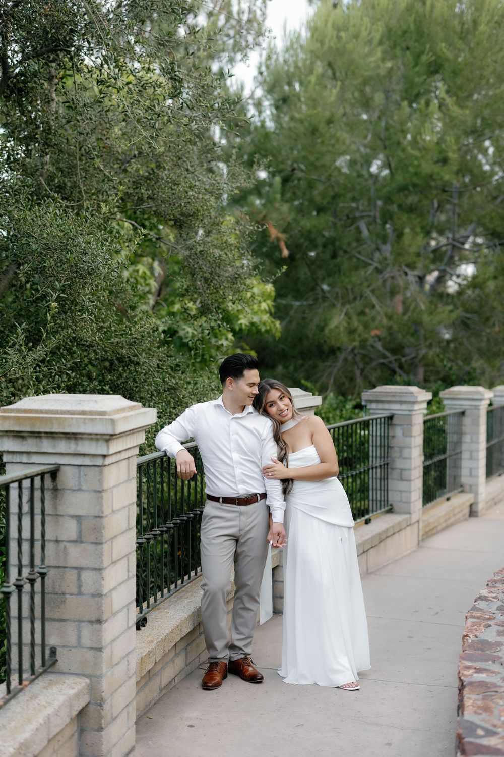 A couple in wedding attire poses on a stone bridge path surrounded by lush green trees.