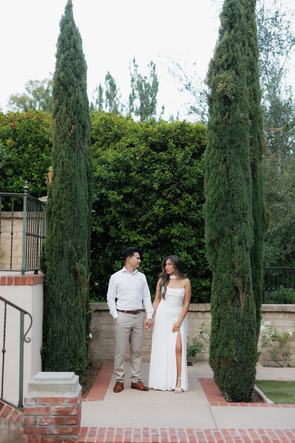Couple in wedding attire holds hands between tall cypress trees on brick pathway in garden.