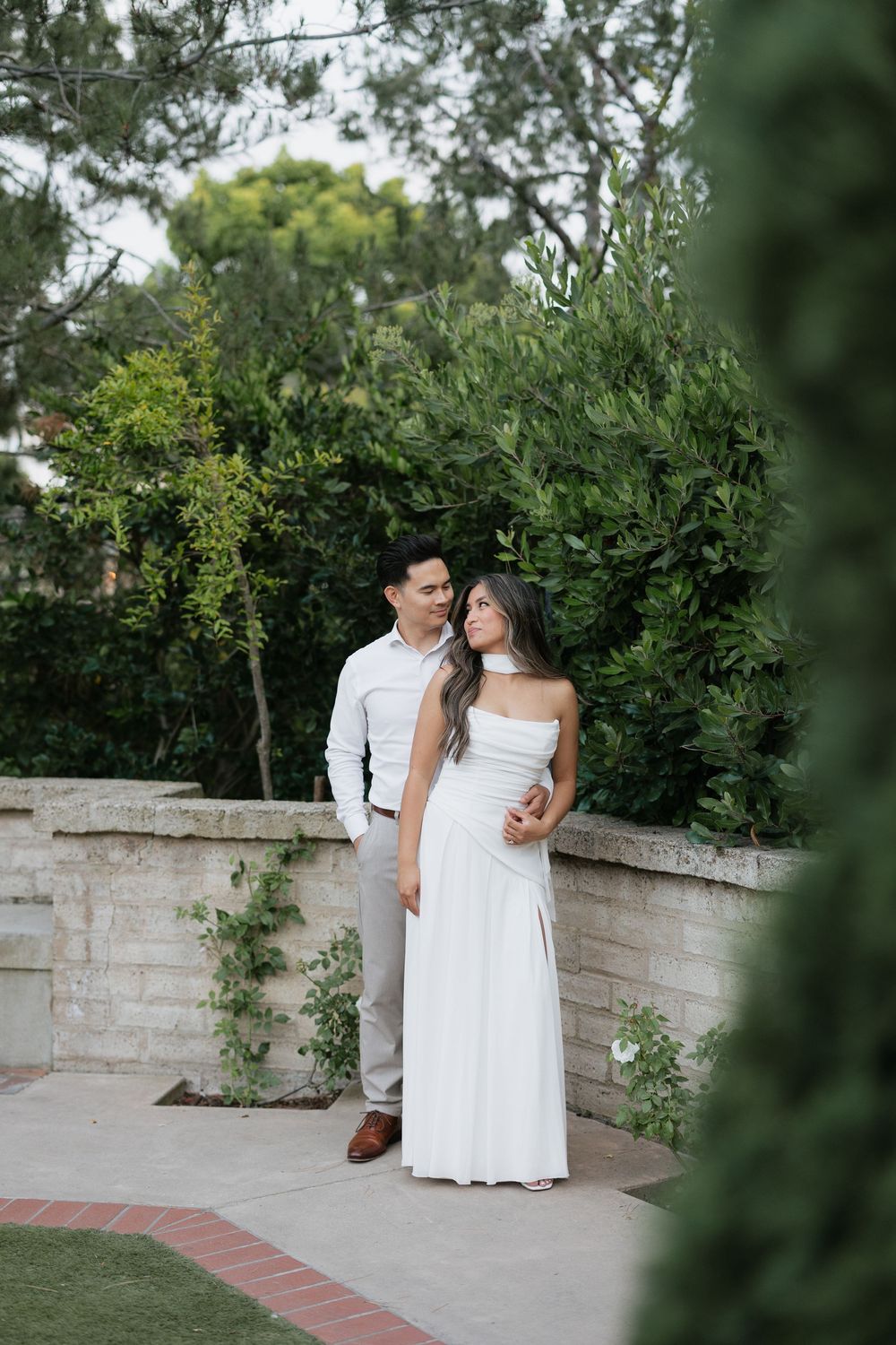 Couple in coordinating white outfits share a romantic moment against a stone wall surrounded by greenery.