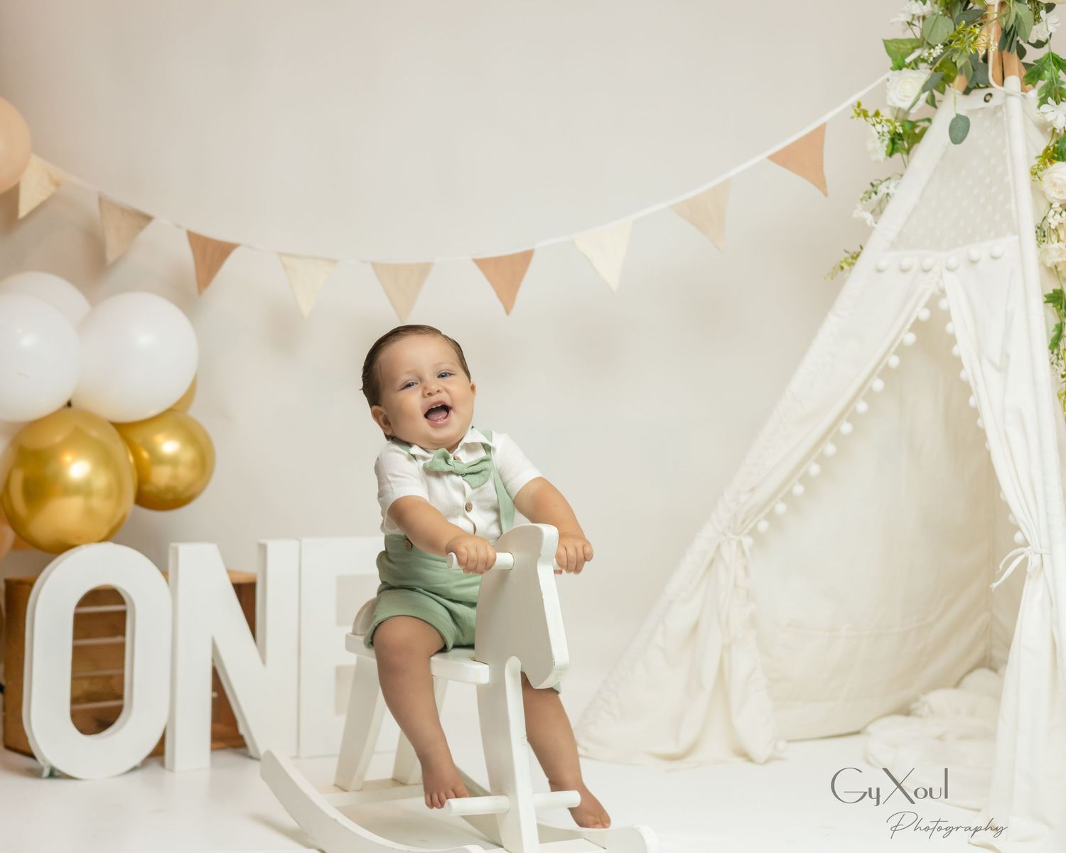 A smiling baby boy in green shorts sits on a white chair surrounded by balloons and a teepee at a first birthday photoshoot.
