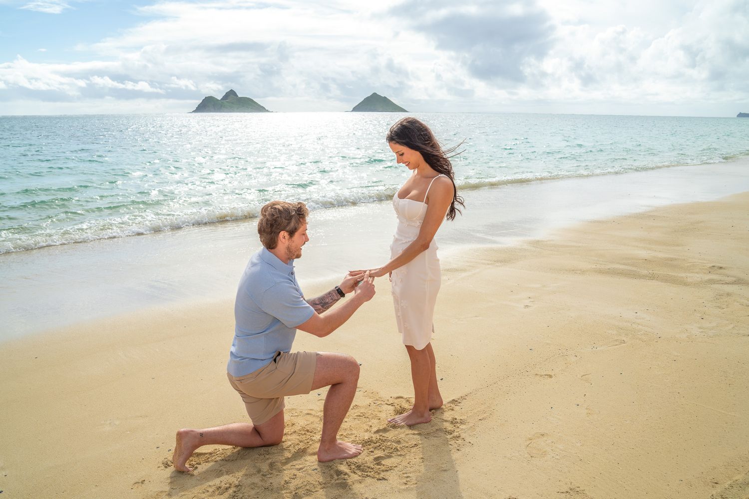 Man proposing to woman on a sandy beach with tropical islands in the background on a sunny day.