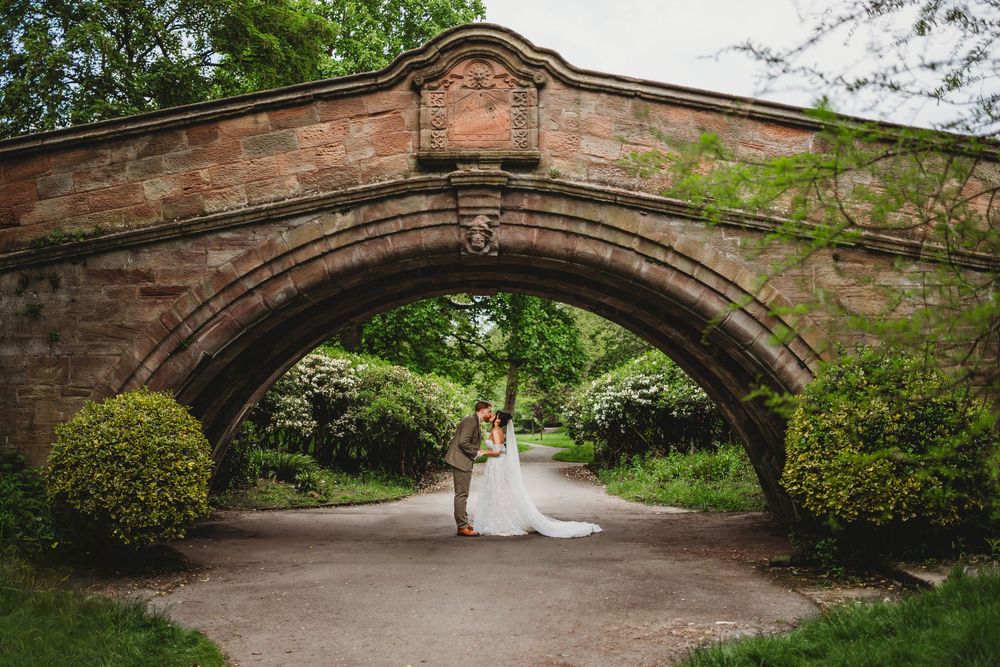 Wedding couple embraces under ornate brick arch bridge surrounded by lush greenery.