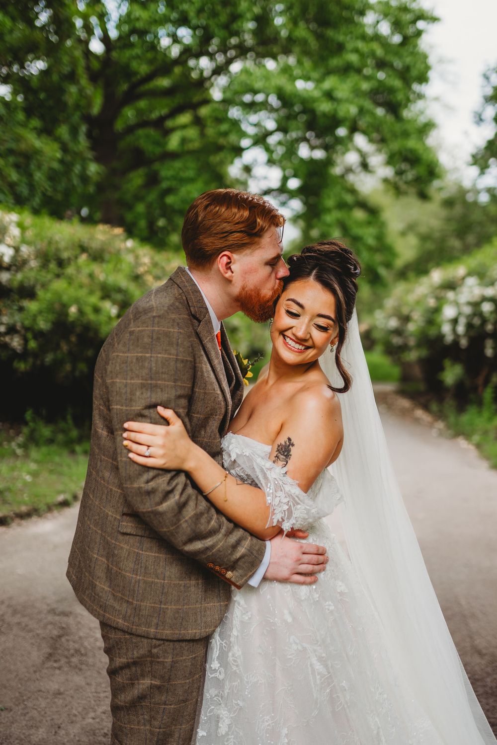 Groom in tweed suit kisses bride's cheek as she smiles in lace wedding dress.