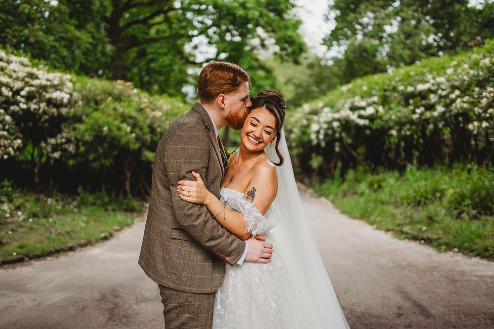 A groom in a tweed suit kisses his bride in a lace wedding dress on a tree-lined path.
