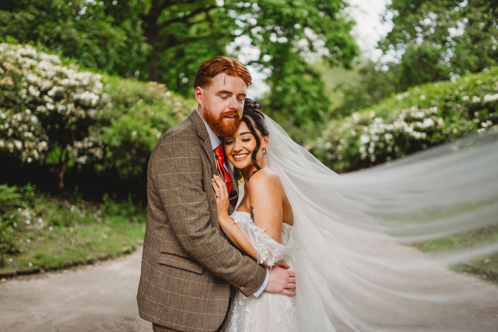 Bride and groom embrace outdoors with flowing veil, surrounded by lush green trees on their wedding day.