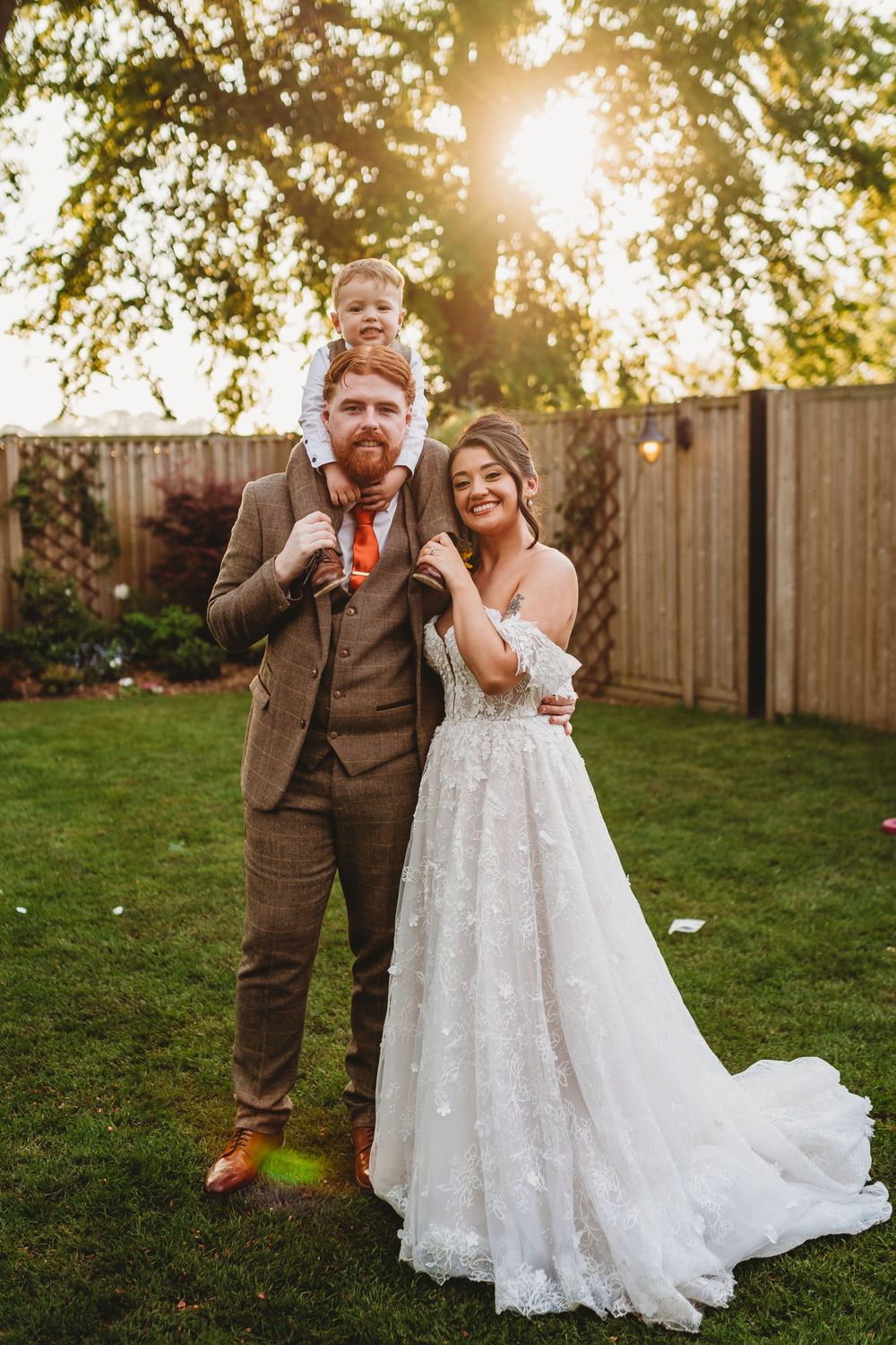 Bride in lace gown and groom in tweed suit pose with young child on shoulders at golden hour wedding.