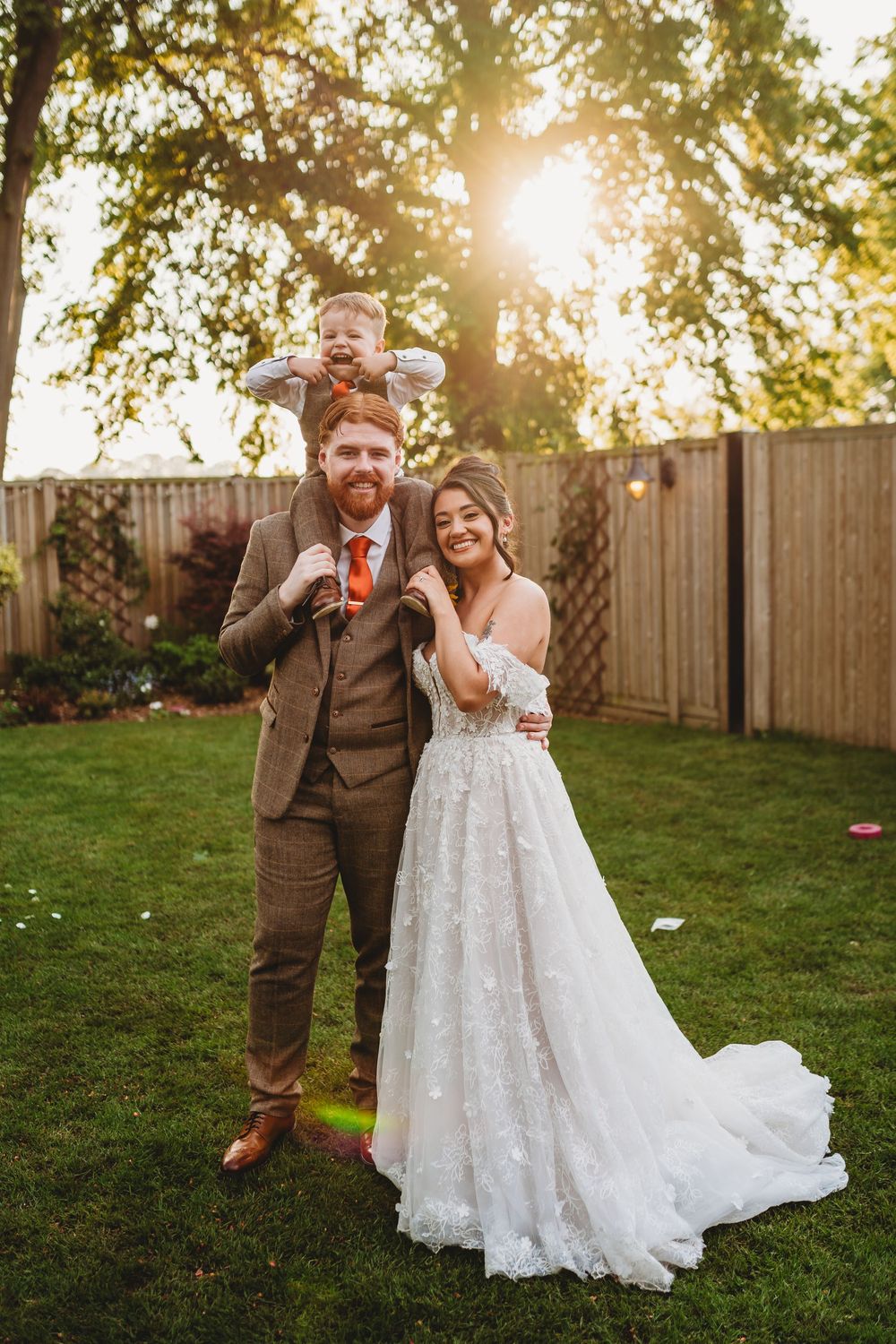Bride and groom pose with young child on shoulders in golden sunset backyard wedding photo.