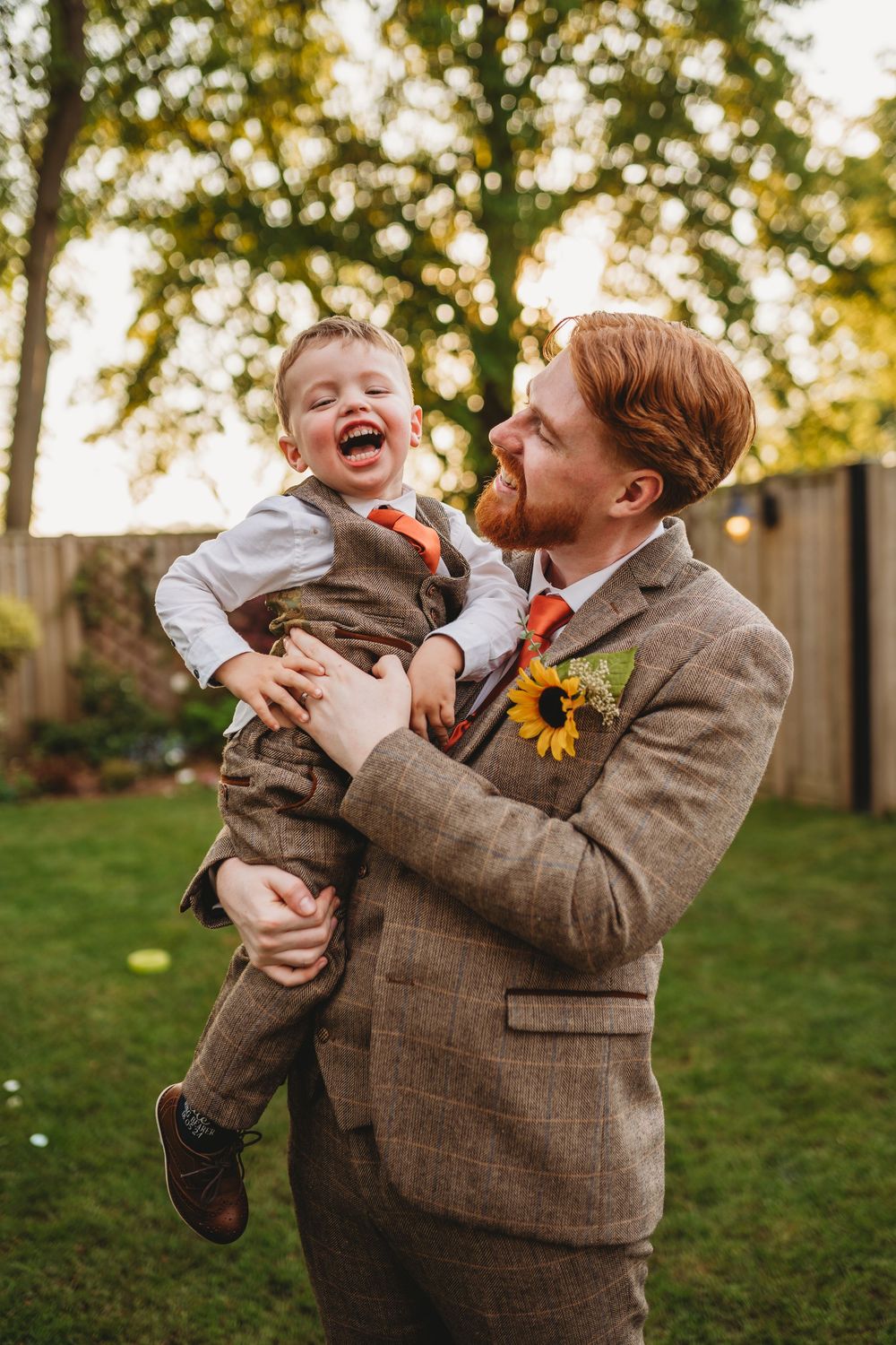 Laughing groom in tweed suit holds giggling young boy in matching outfit at outdoor wedding reception.