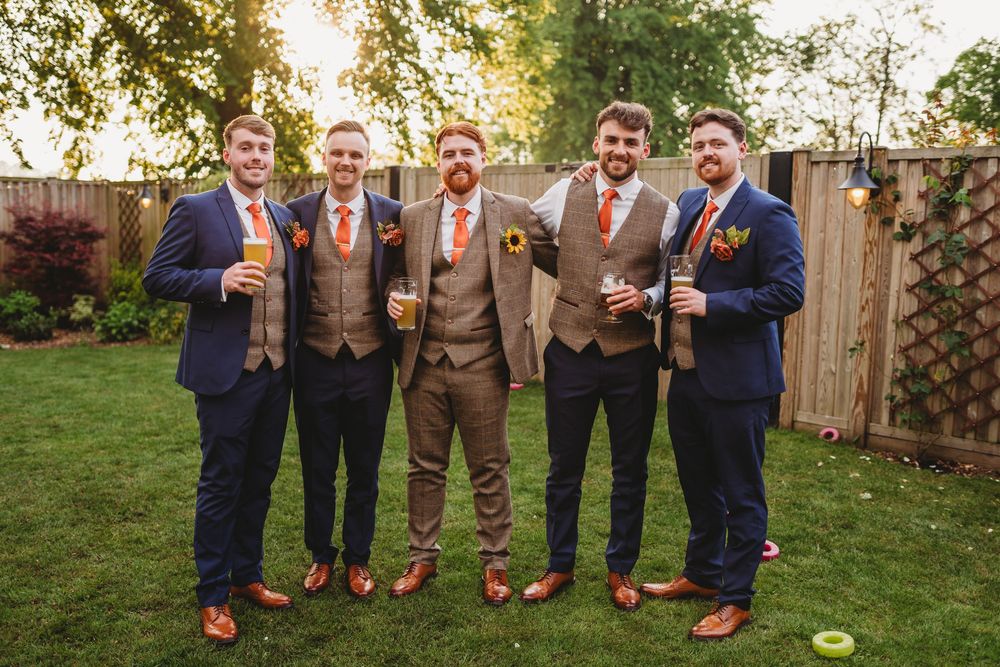Groom and groomsmen in navy and brown suits with orange ties posing outdoors at a garden wedding.