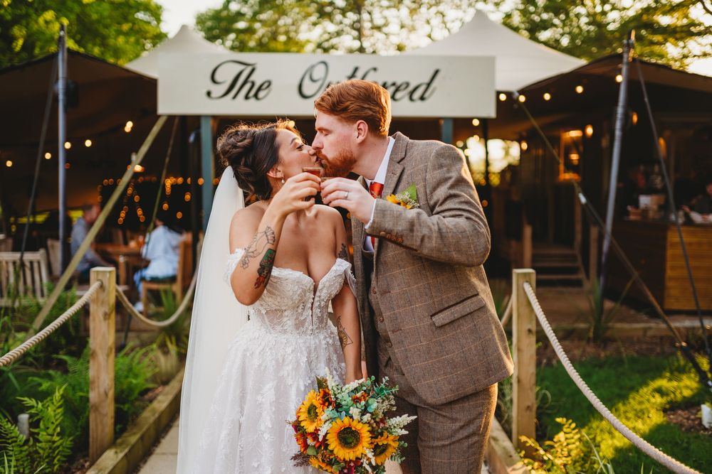 Bride and groom share a kiss outdoors at The Orchard venue, bride holding sunflower bouquet in lace gown.