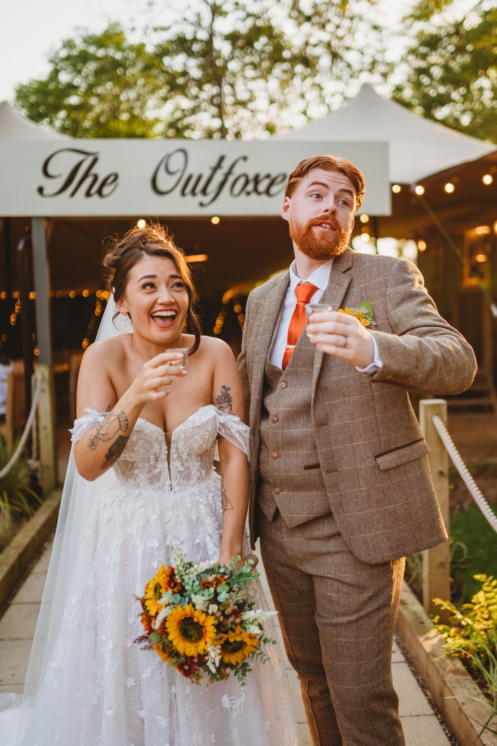 A joyful bride with sunflowers and tattooed groom in tweed suit celebrate outside The Outfoxed venue.