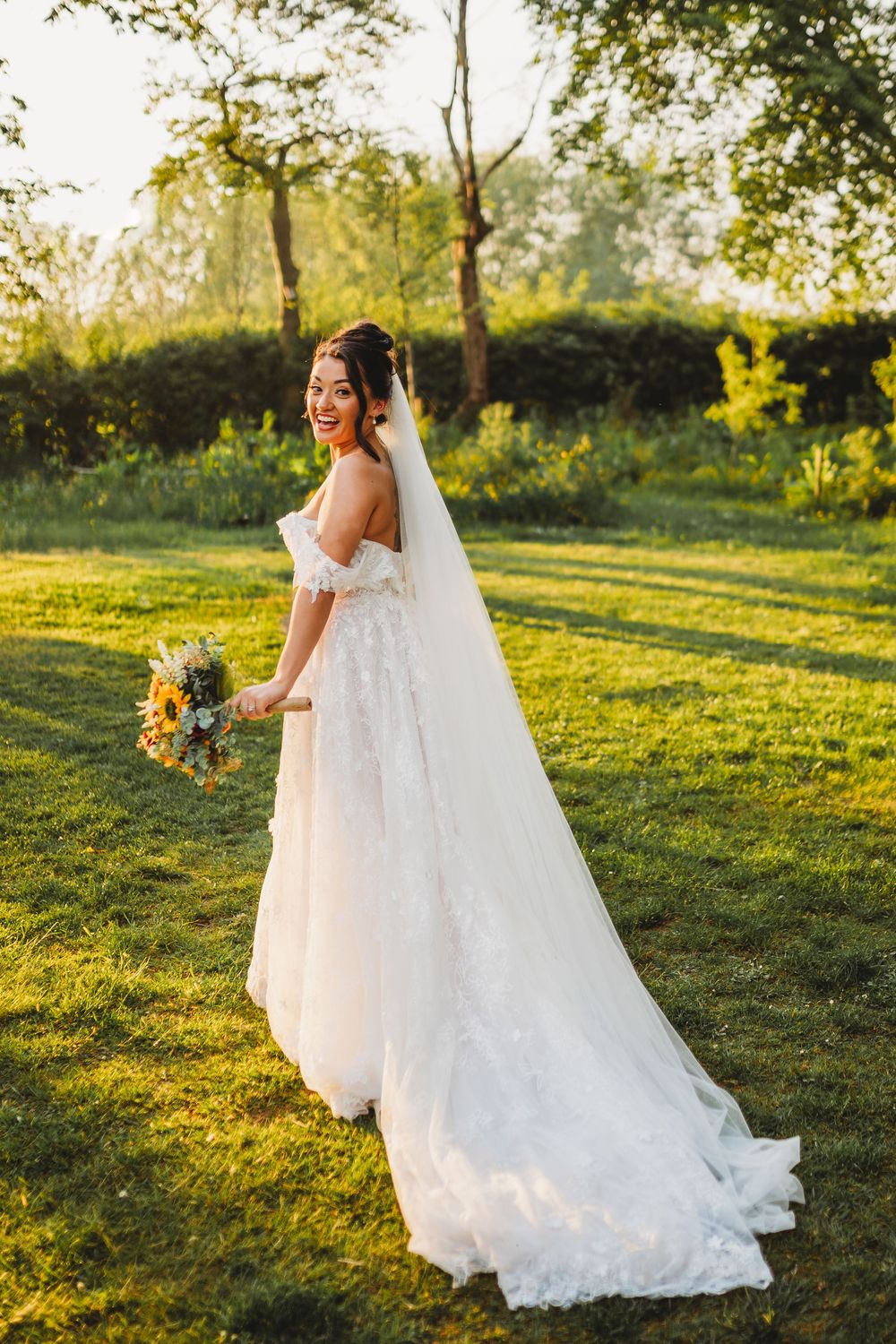 Smiling bride in white lace gown with veil holds bouquet on sunlit green lawn.