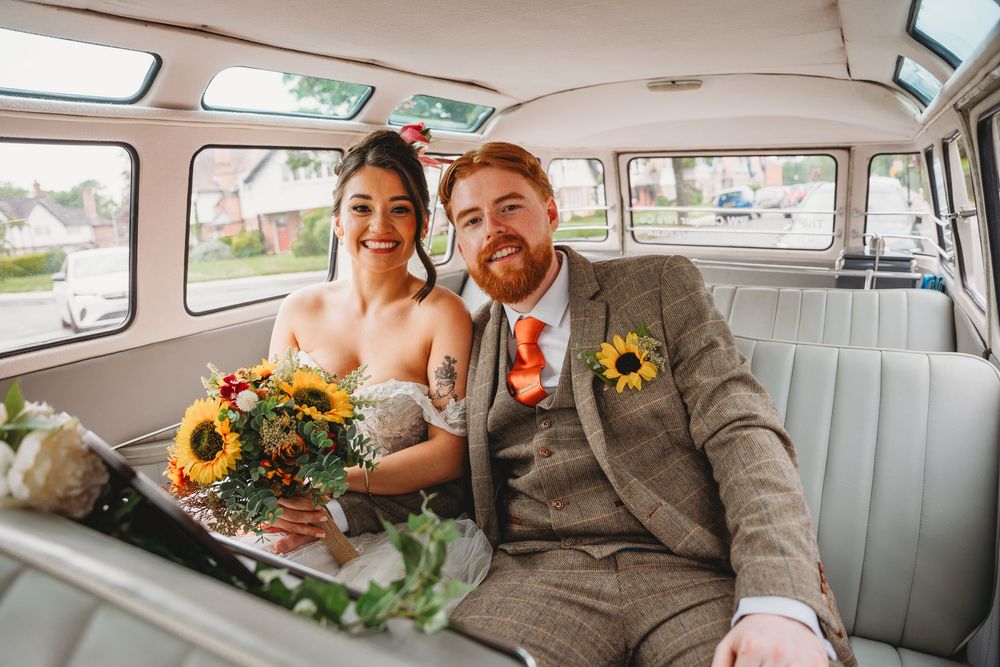 Smiling bride and groom sit inside vintage white bus, bride holding sunflower bouquet on wedding day.