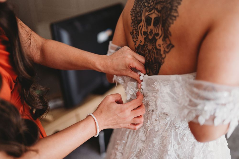 Bridesmaid helping bride button up lace wedding dress, bride has large back tattoo.