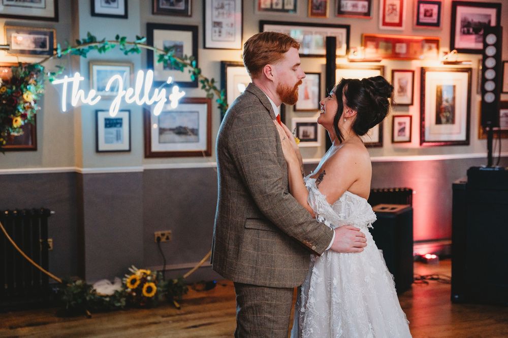 Bride and groom share first dance under neon sign reading The Jellys at rustic wedding venue with gallery wall.