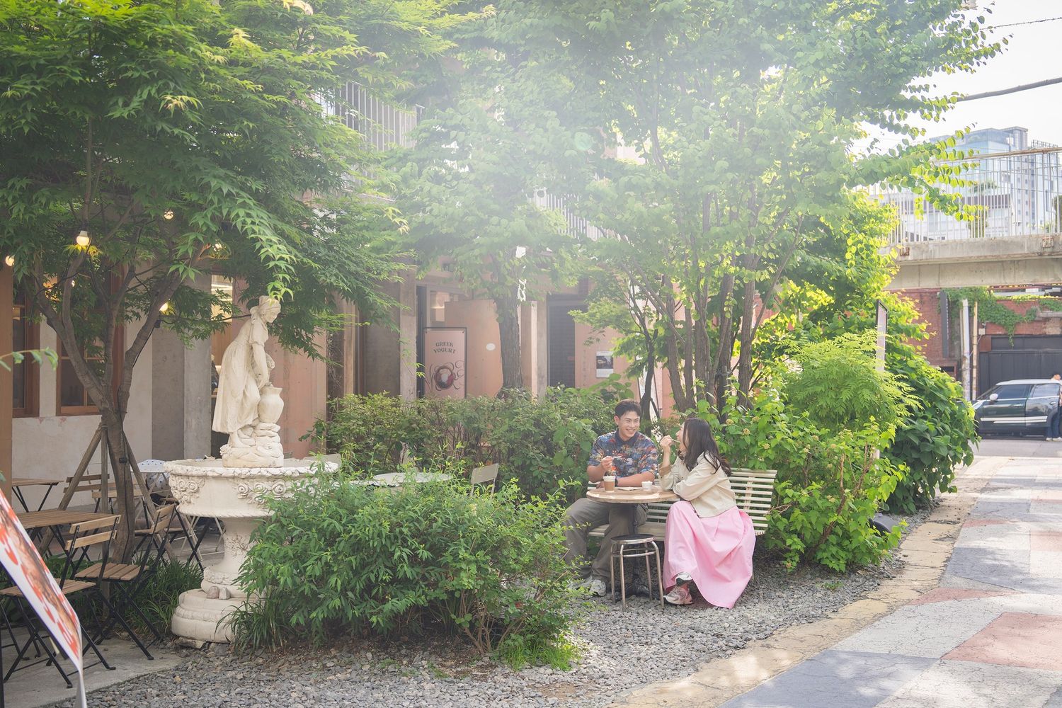 Couple sitting outdoors in a lush garden cafe with a classic statue fountain surrounded by green trees.
