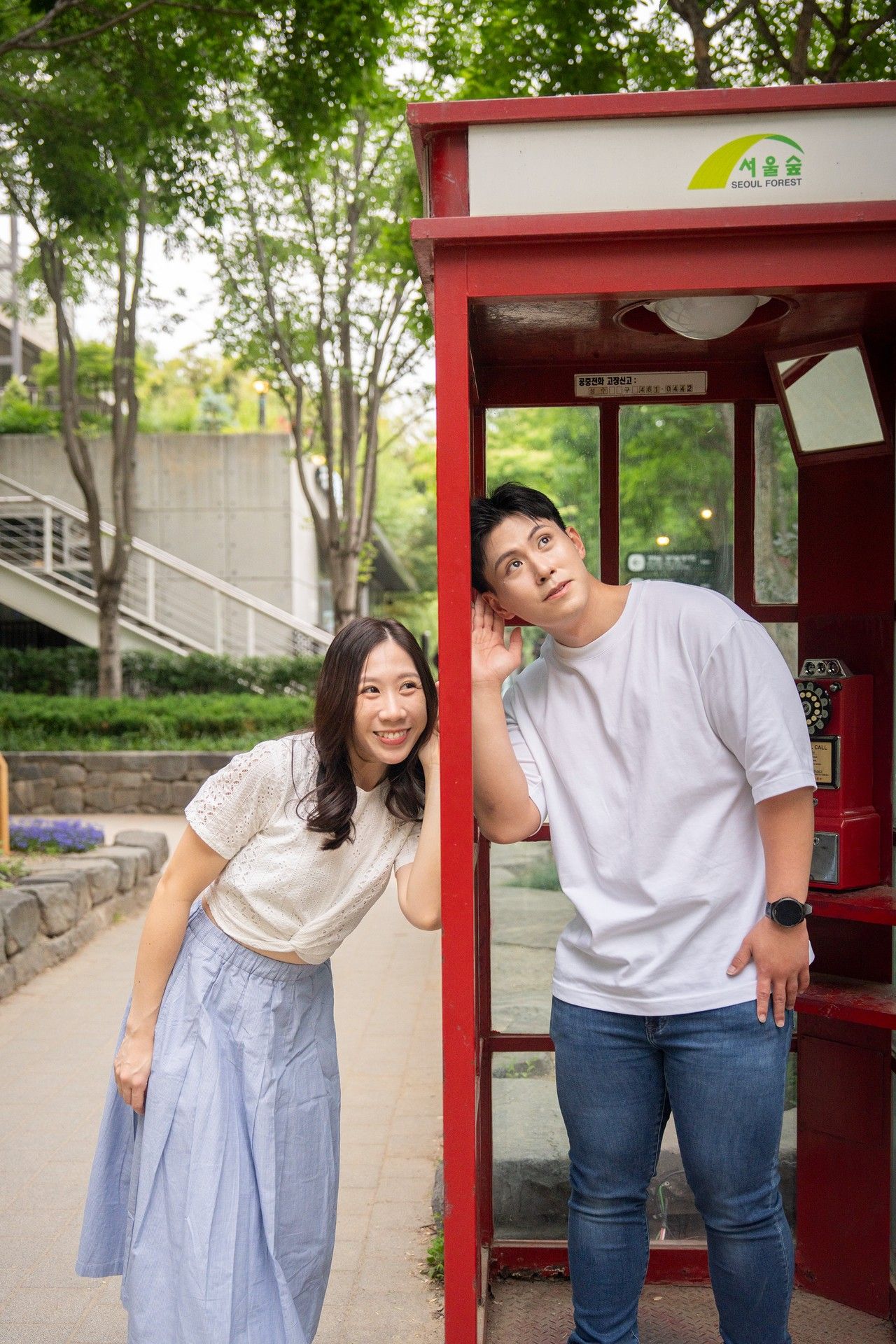 Young Asian couple posing by a red phone booth outdoors, smiling in casual summer outfits near a tree-lined path.