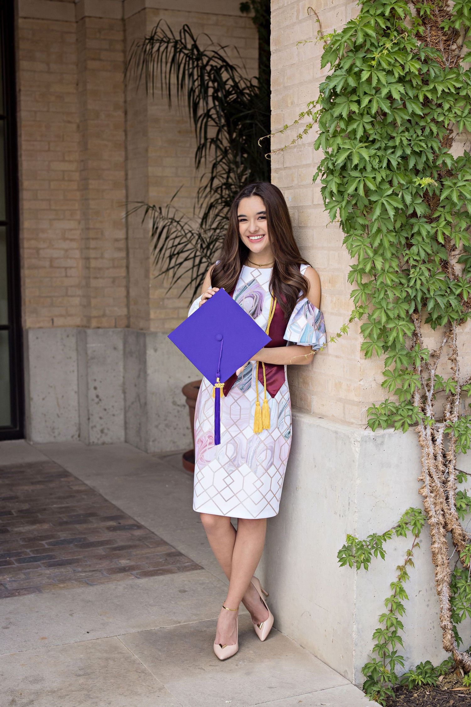 Smiling young woman in purple graduation cap and gown with gold cords and maroon stole, class of 2022.