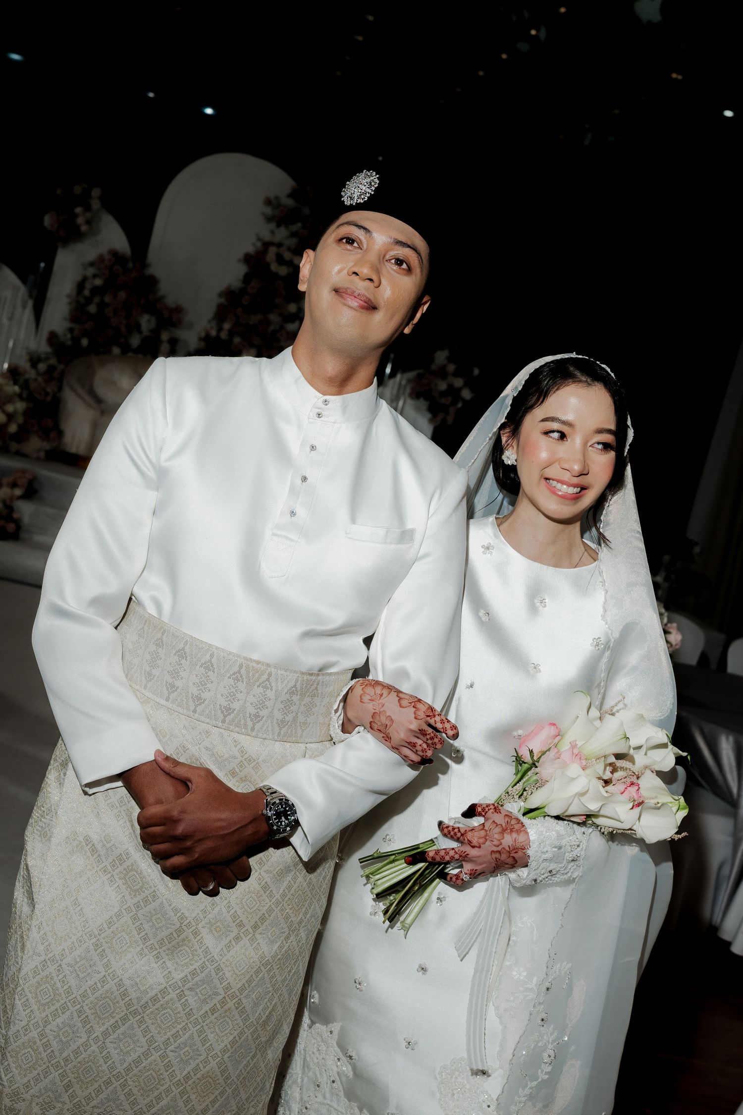 Malay couple in traditional white wedding attire, groom in baju melayu, bride holding pink and white bouquet.
