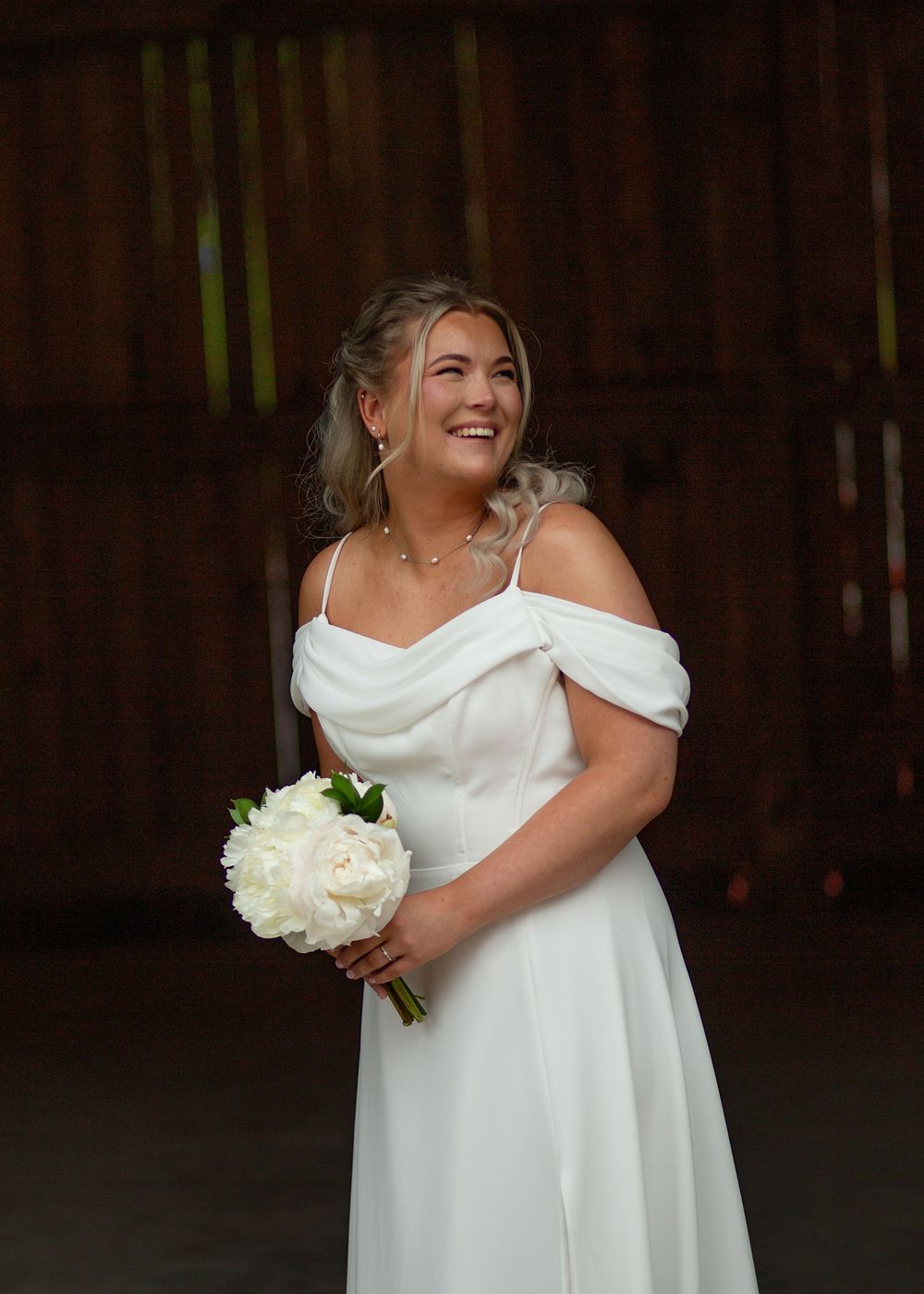 Smiling bride in white off-shoulder gown holding white rose bouquet, standing in rustic barn setting.