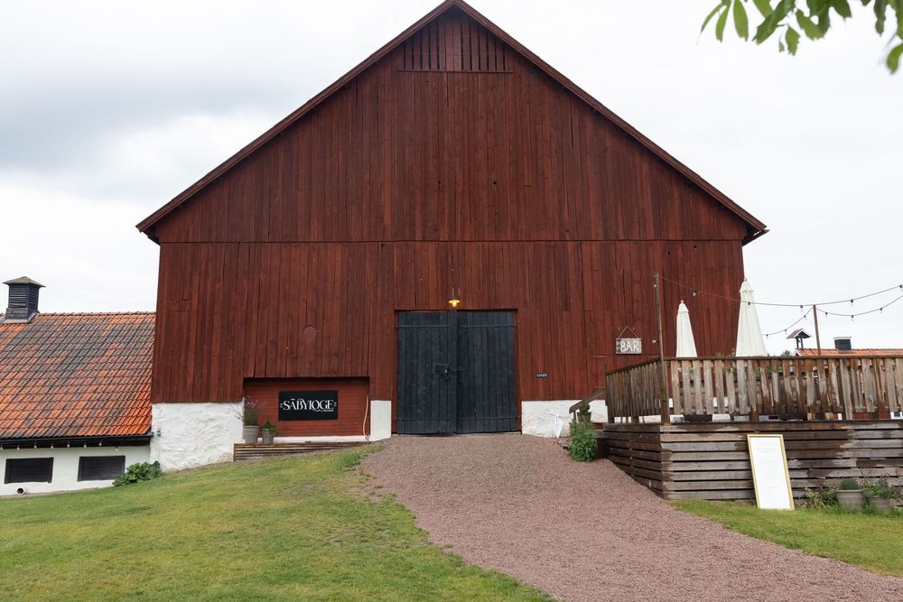 Red wooden barn with dark double doors and a small sign, set on a green lawn under an overcast sky in rural Sweden.