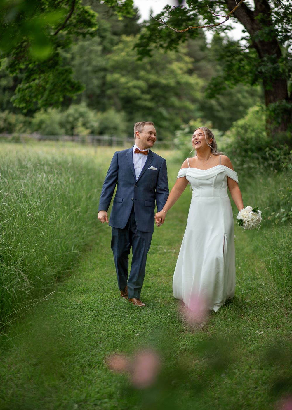 A joyful bride and groom walk hand-in-hand through a lush green meadow path on their wedding day.