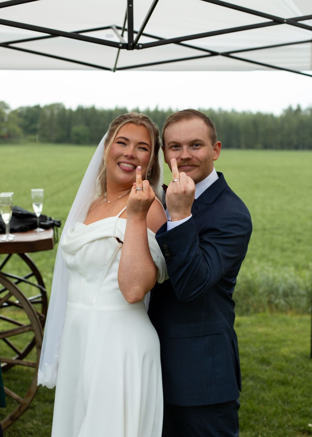 Newlywed couple in wedding attire showing off their wedding rings outdoors on a green field.