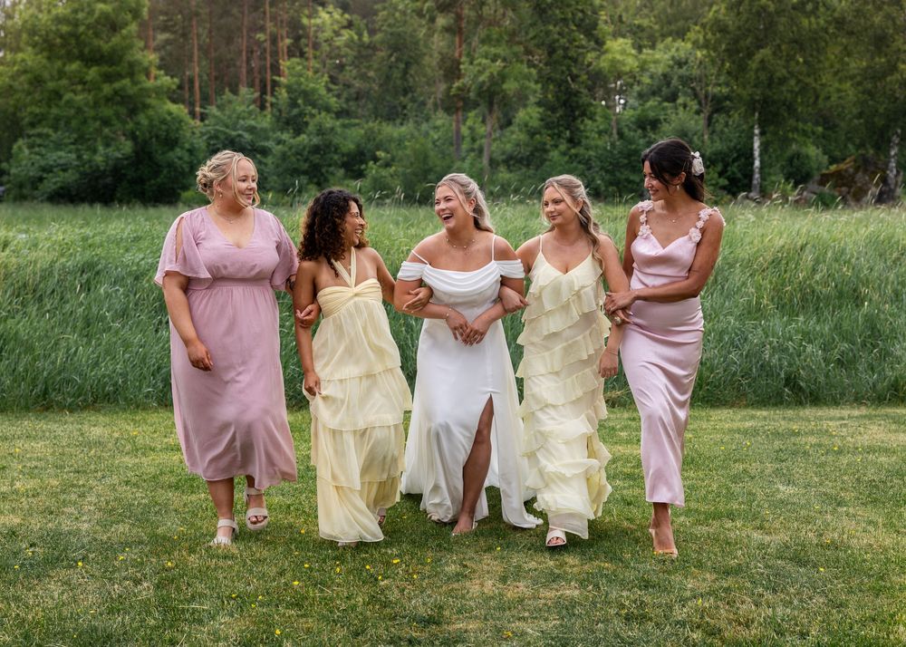 Bride in white gown walks with four bridesmaids in pink and yellow dresses on a green lawn.