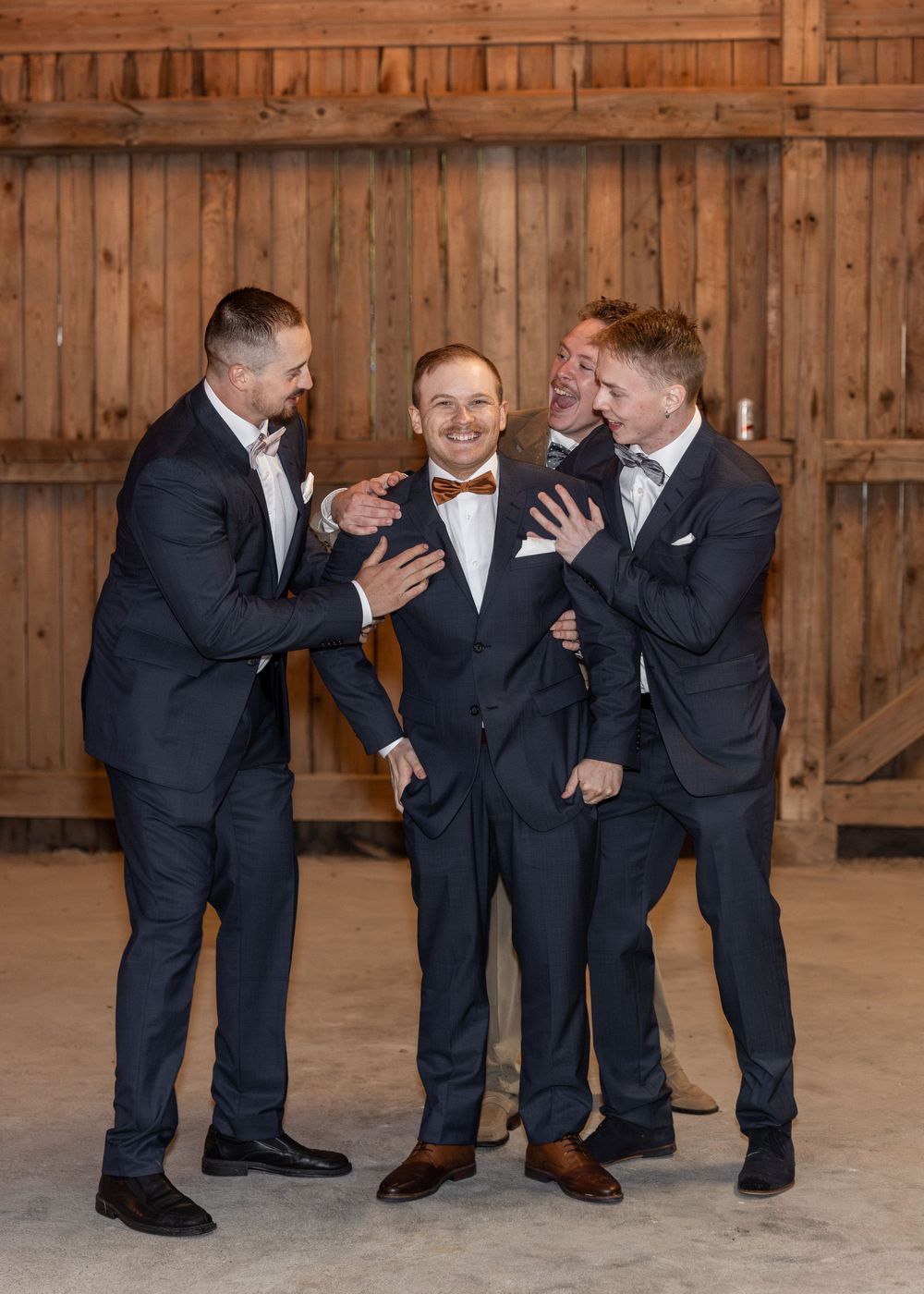 Groom in navy suit and bow tie laughing with groomsmen in matching suits inside a rustic barn.