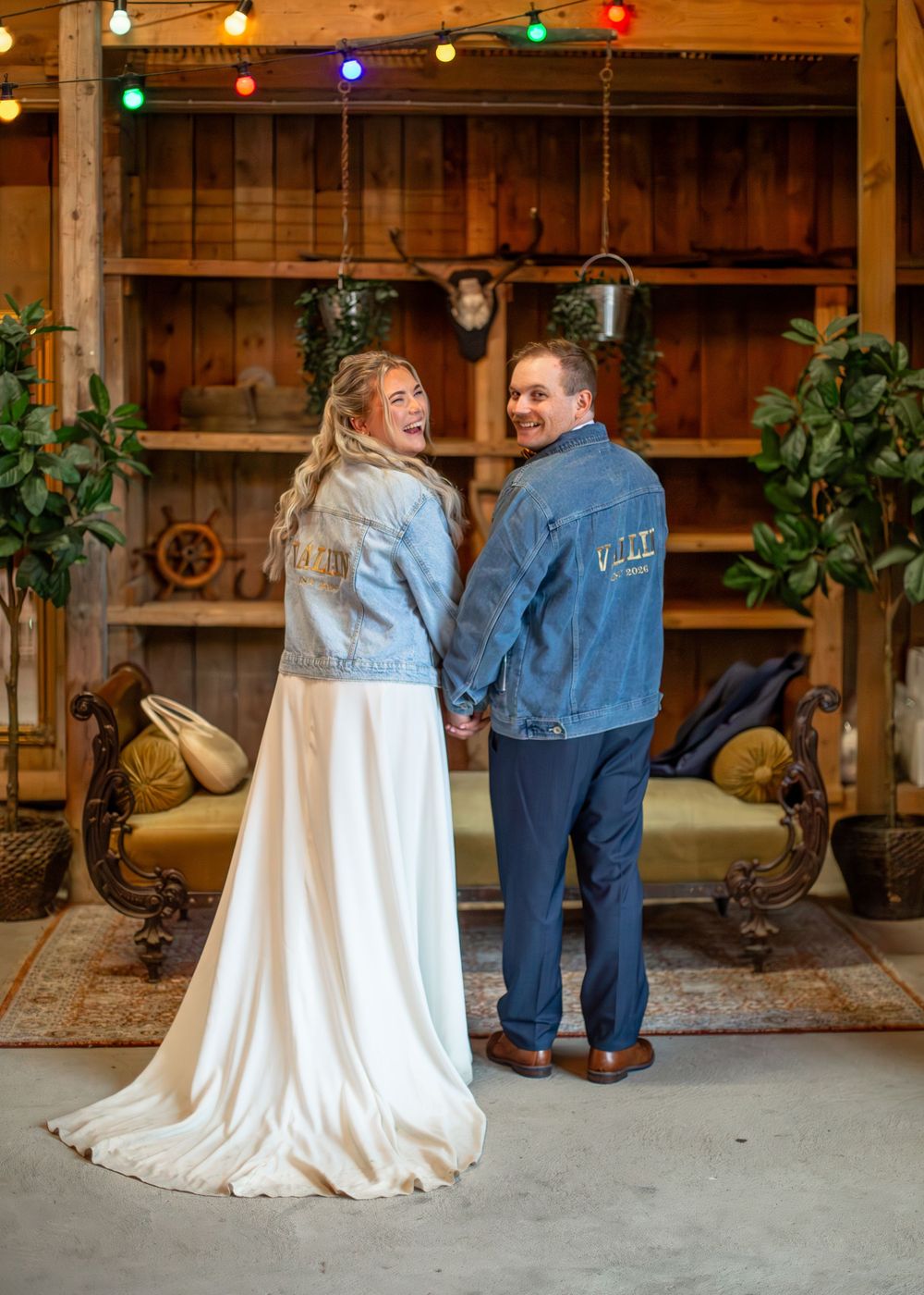 A smiling bride and groom in matching denim jackets hold hands inside a rustic barn wedding venue.