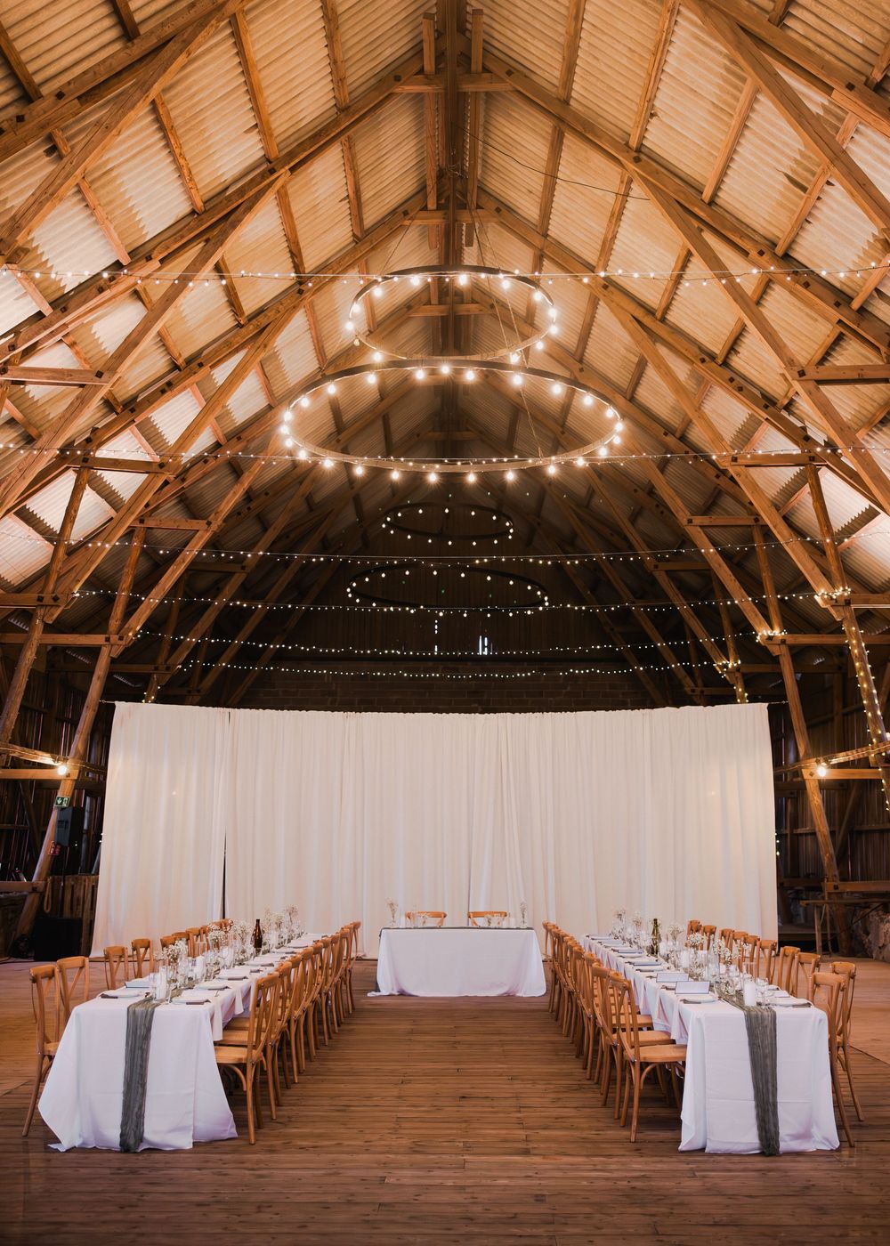 Elegant barn wedding reception with string lights, long tables, and white drape backdrop.