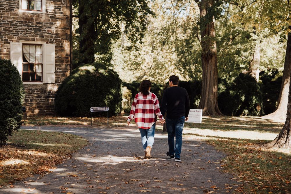 Couple walking hand in hand along a tree-lined path beside a stone building in autumn.
