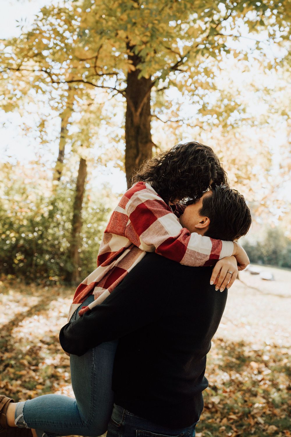 Couple embracing and kissing in autumn park, woman in red plaid shirt lifted by partner among golden fall foliage.