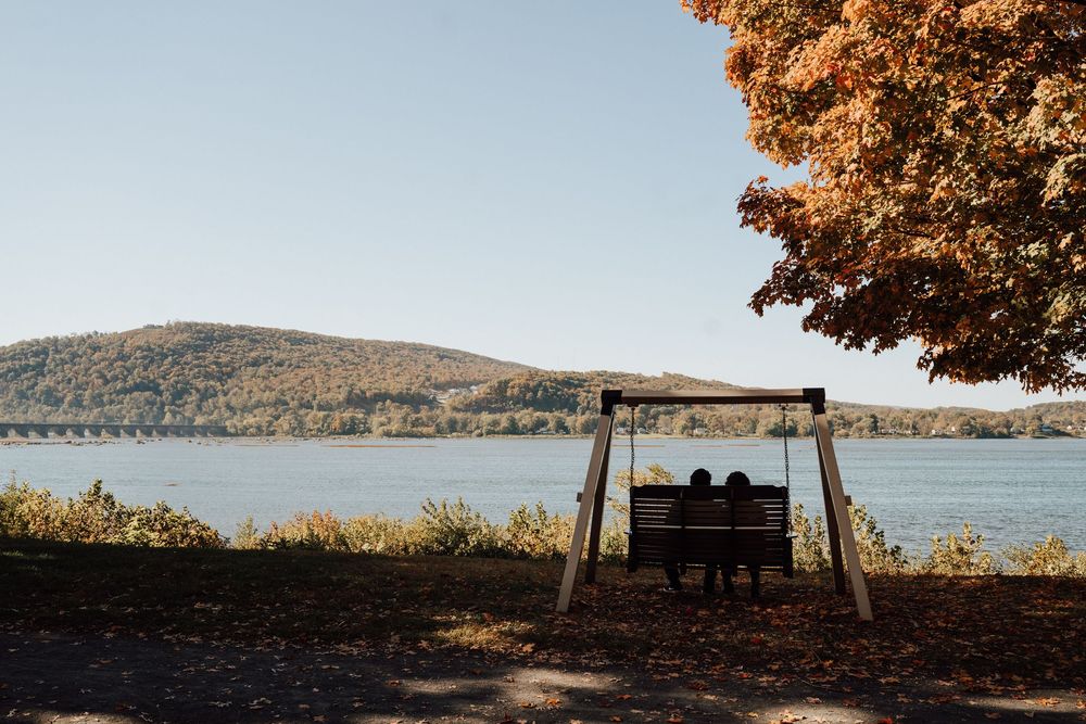 Couple sitting on a wooden swing bench overlooking a serene lake with autumn foliage and rolling hills in the background.