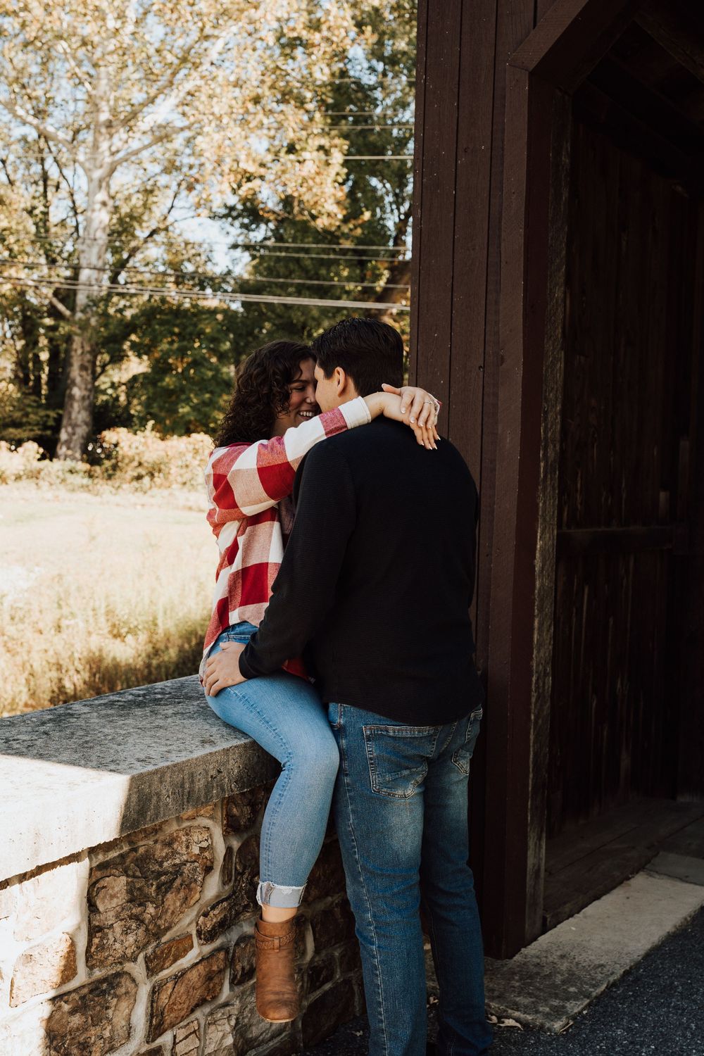 Couple sharing a romantic embrace near a rustic stone wall and barn in autumn setting.