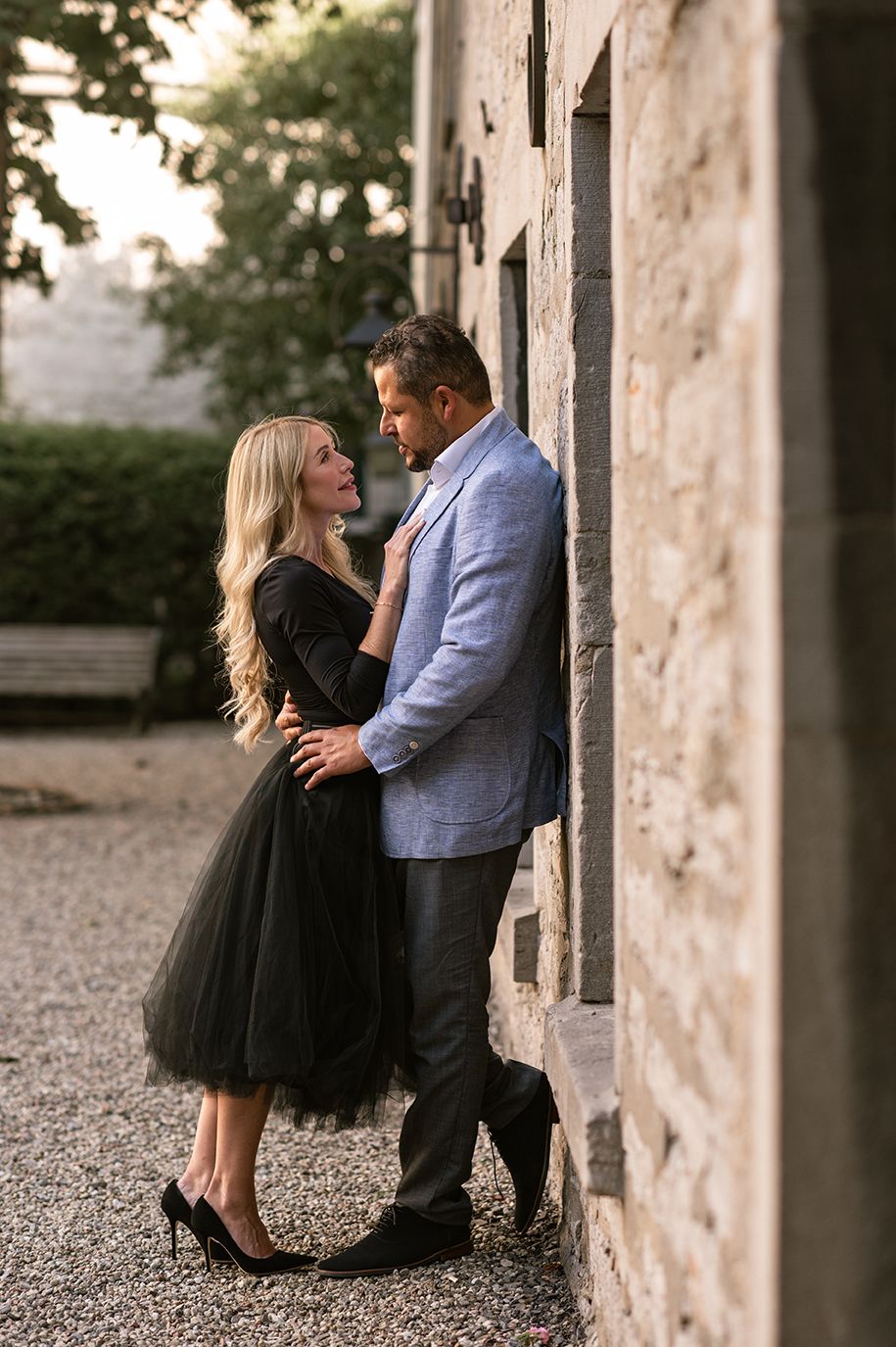 Couple in elegant attire sharing a romantic moment against a stone wall outdoors.