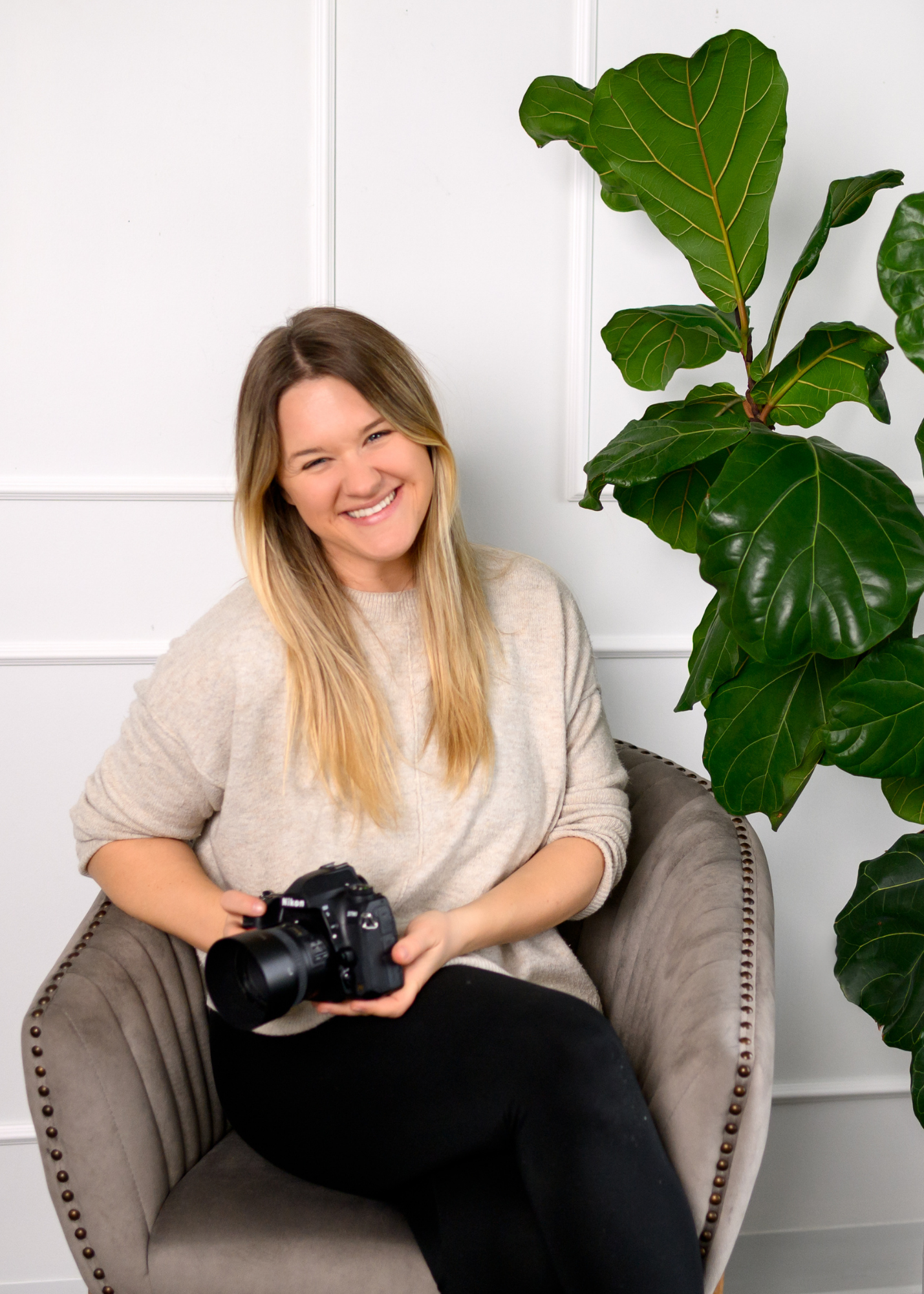 Smiling woman photographer sitting in gray chair holding DSLR camera next to fiddle leaf fig plant in bright white studio.