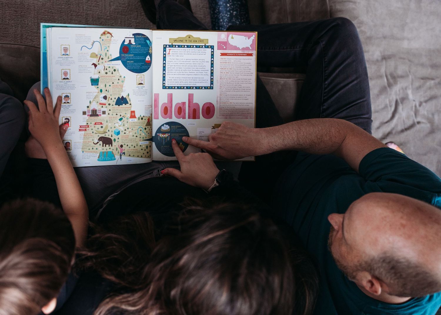 Adult and two children sit on a couch reading a colorful illustrated map book together.