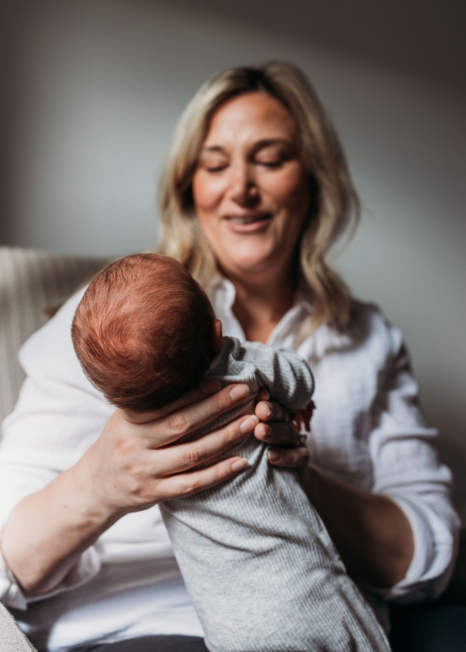 A smiling blonde mother tenderly holds her newborn baby with red hair in her arms while seated in a chair.