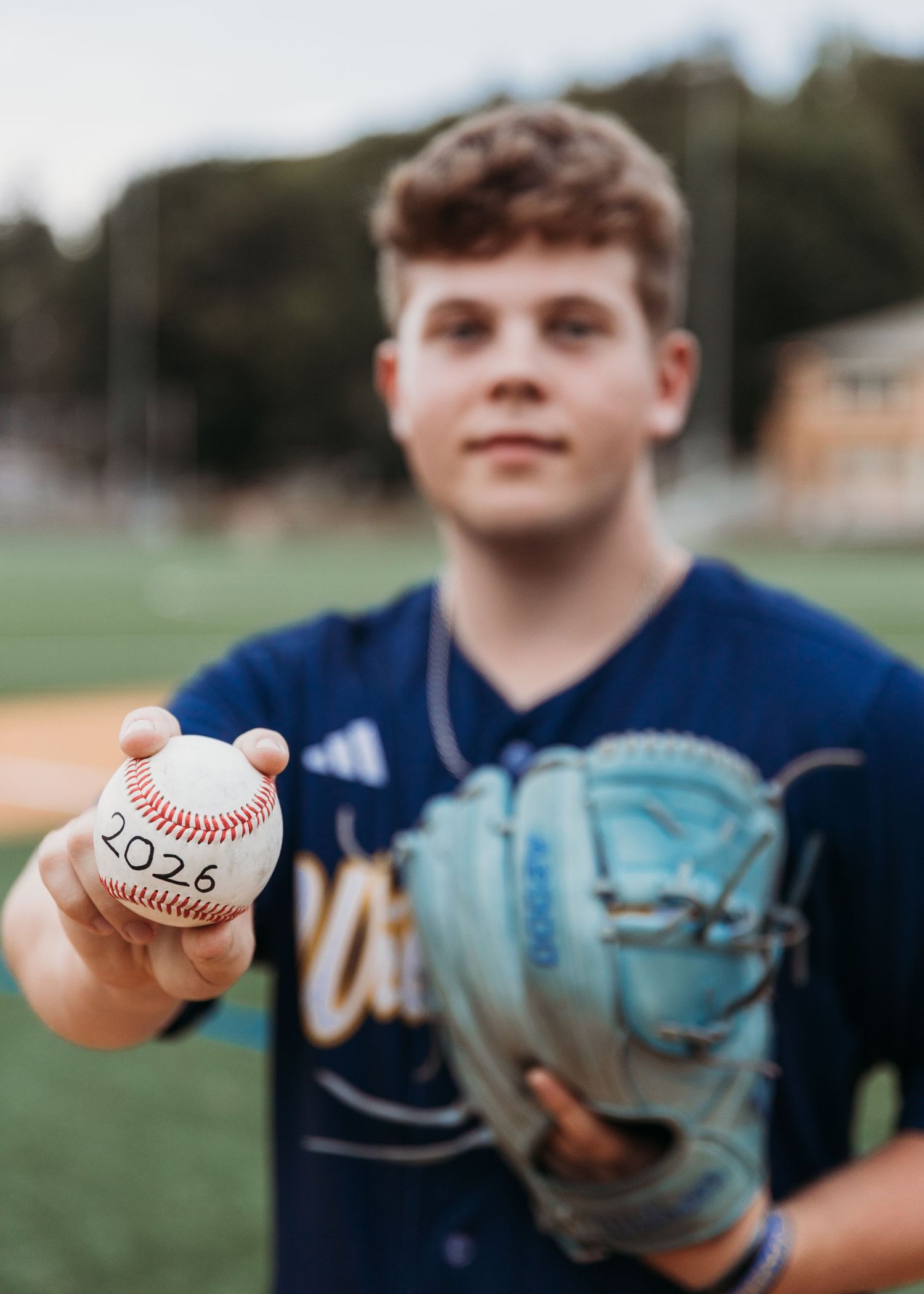 A teenage baseball player holds a ball marked '2026' toward the camera while holding a glove on a field.