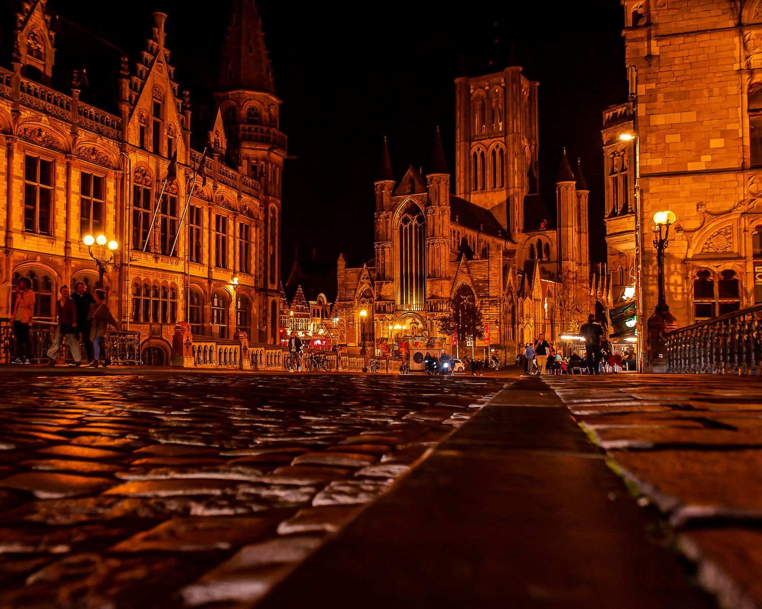 Wide nighttime photo of a brightly lit city square with historic buildings and street lights, reflecting off wet pavement. Gent Belgium
