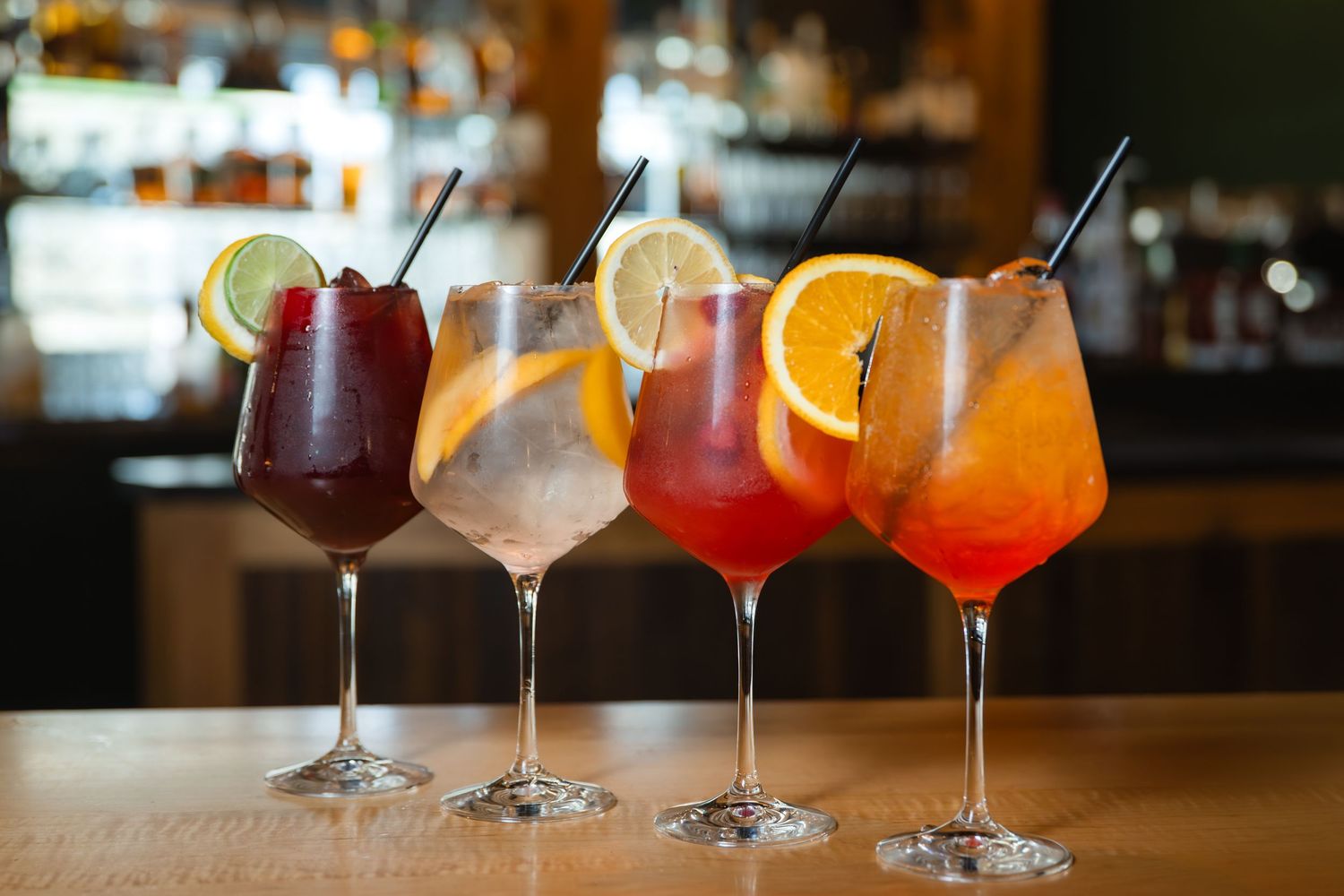 Four colorful cocktails in wine glasses with fruit garnishes and black straws lined up on a wooden bar counter.