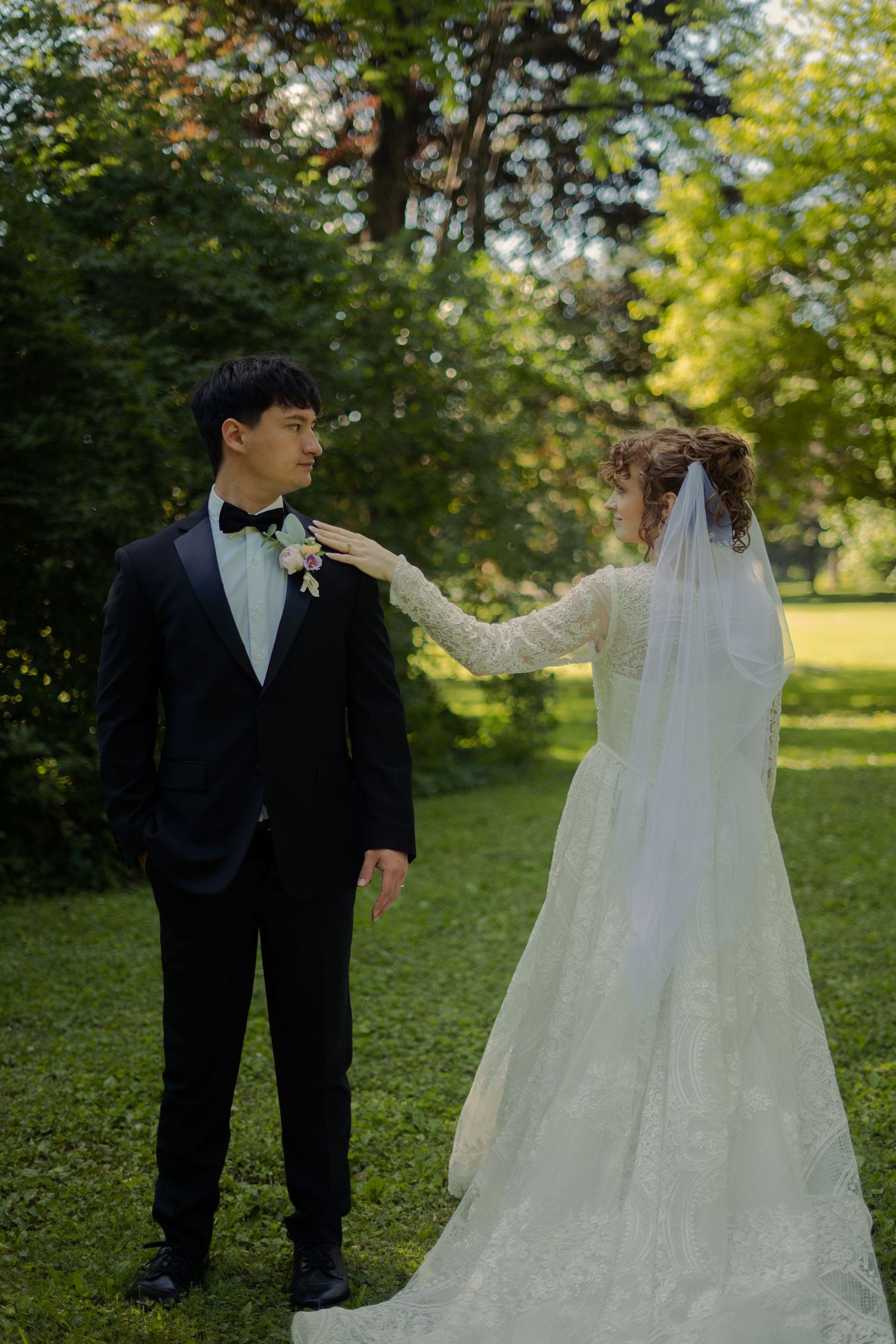 Bride in lace gown touches grooms lapel as they stand on green lawn in sunlight.