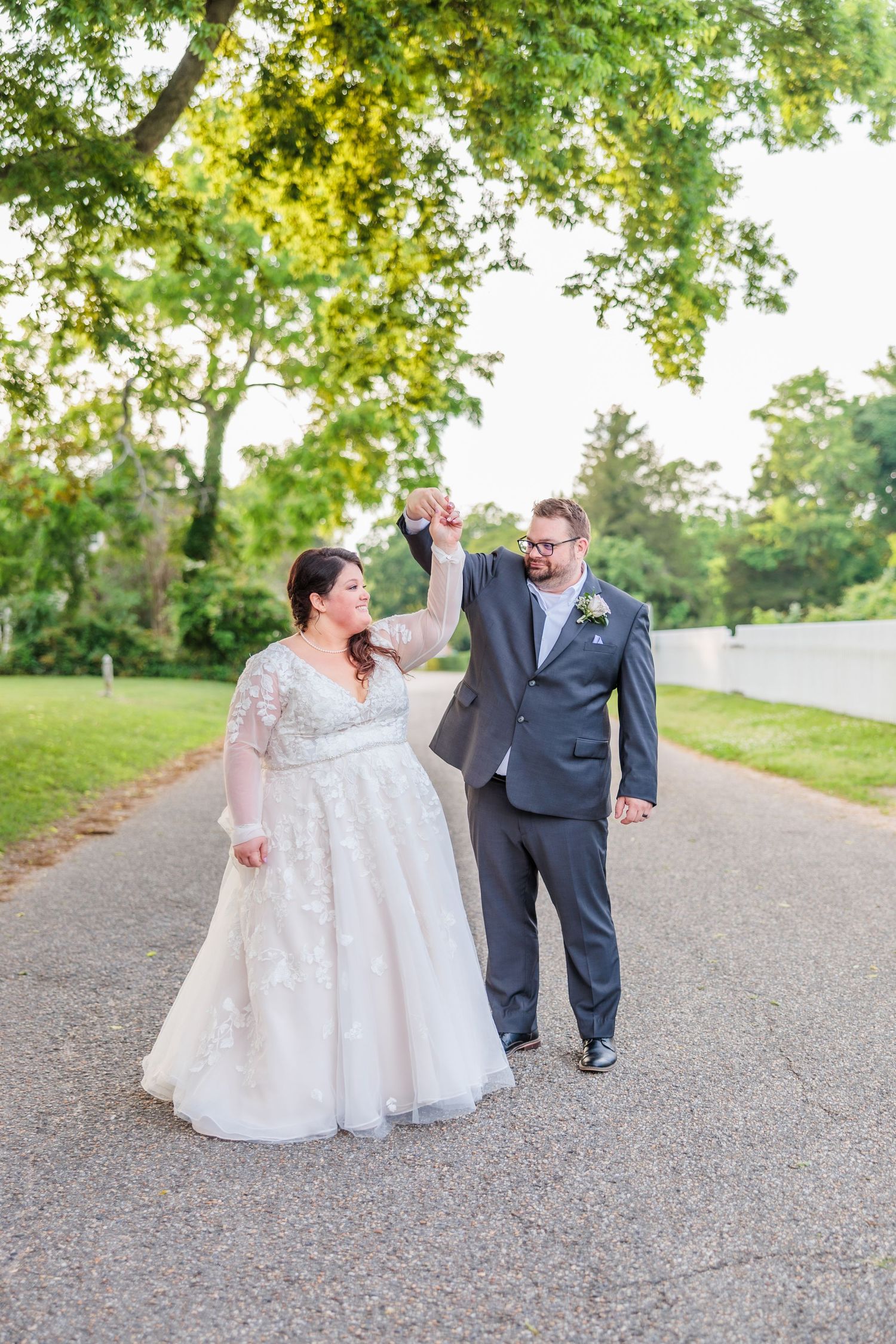 A couple celebrates their wedding day on a tree-lined path with a joyful dance move.