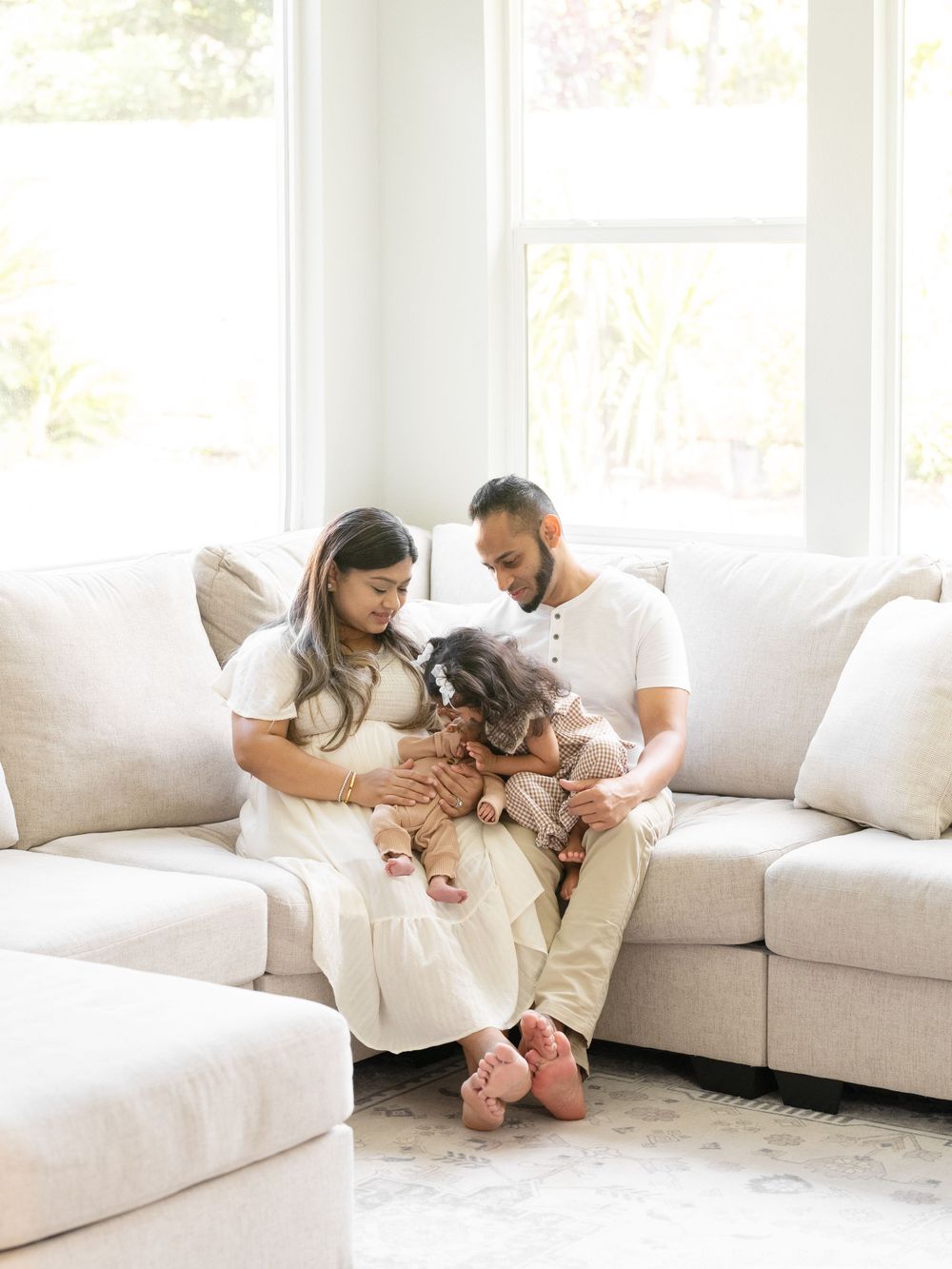 A young family sits together on a cream sofa, parents lovingly gazing at their toddler in a bright living room.