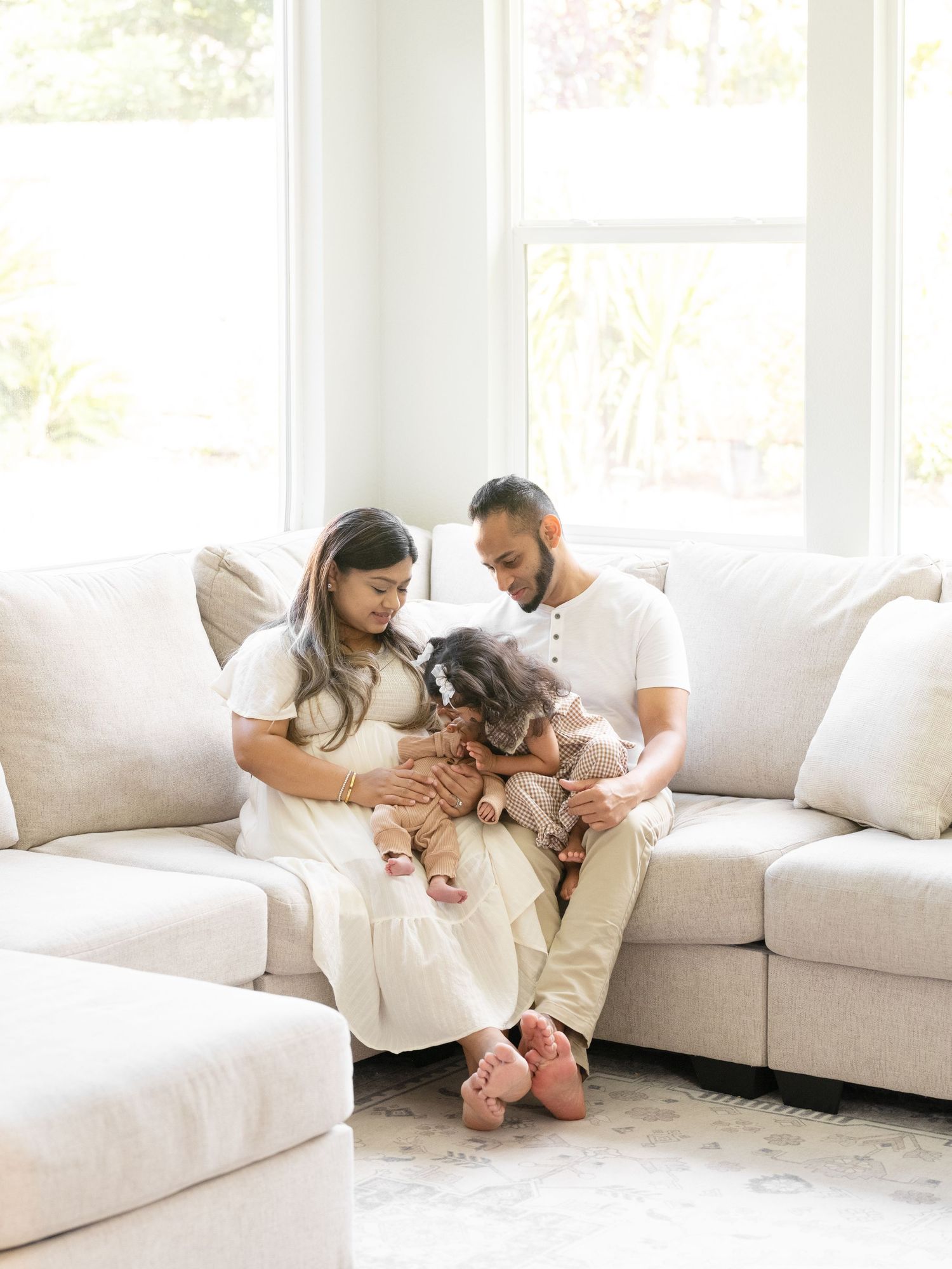 A young family sits together on a cream sofa, parents lovingly gazing at their toddler in a bright living room.