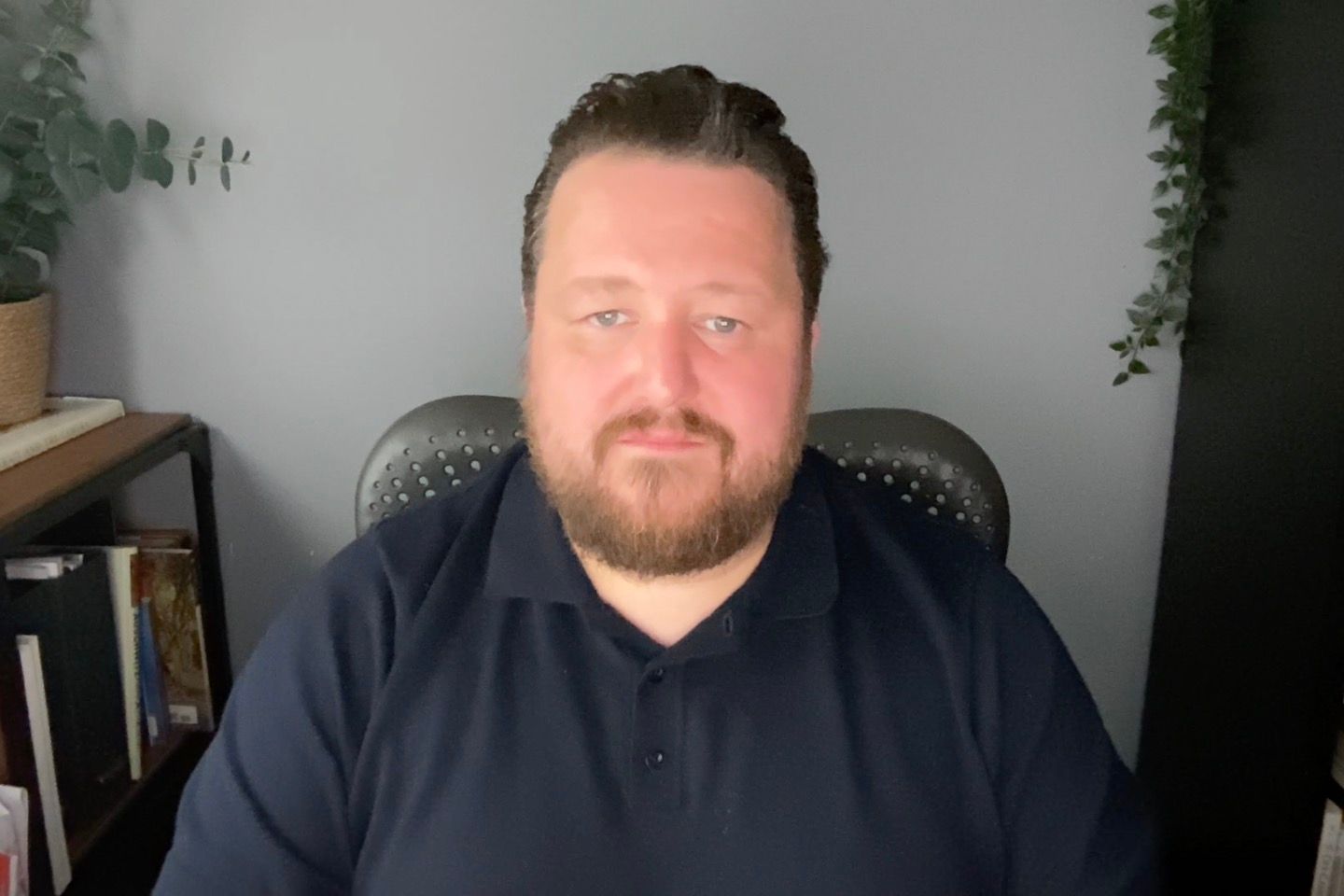 A man with a beard in a navy polo shirt sits in a home office chair against a gray wall with plants.