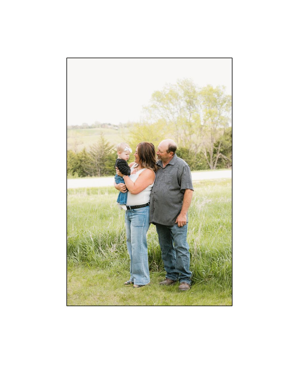 A couple stands together in a green field, lovingly gazing at their baby on a sunny day.
